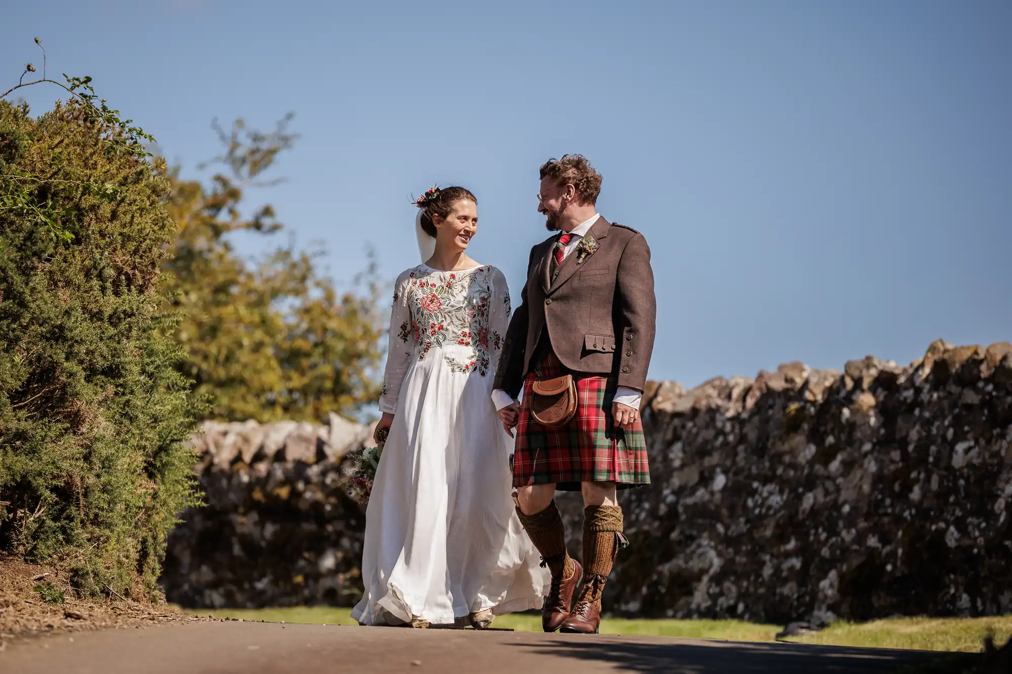 A bride in a long white floral dress and a groom in a brown jacket, red tartan kilt, and sporran walk hand-in-hand outdoors on a sunny day, smiling at each other, with stone walls and greenery in the background.