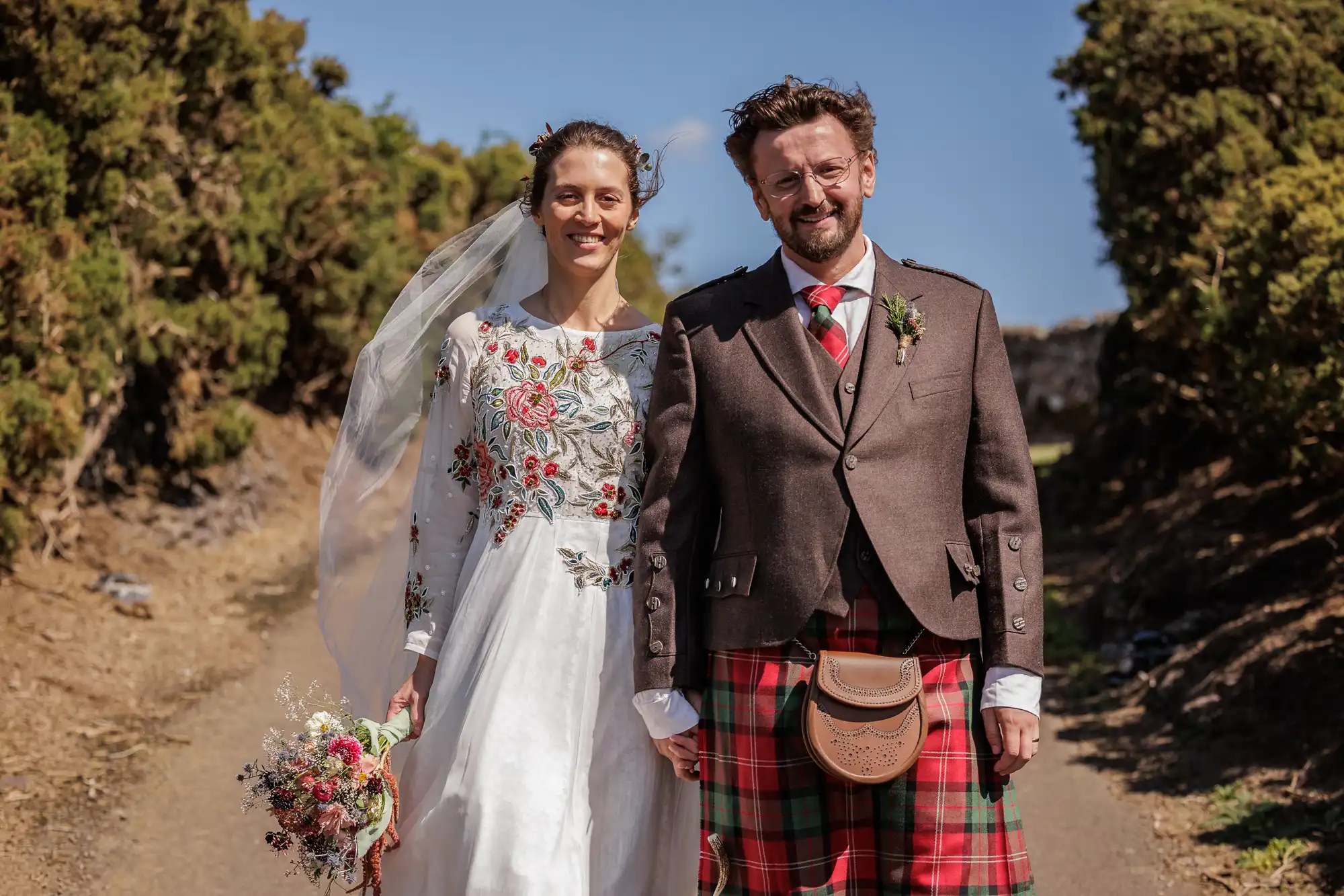 A smiling bride in a white floral-embroidered dress stands beside a groom in a brown jacket, red tartan kilt, and sporran. They hold hands on a sunlit path lined with green bushes; the bride holds a bouquet and wears a long veil, under a clear blue sky.