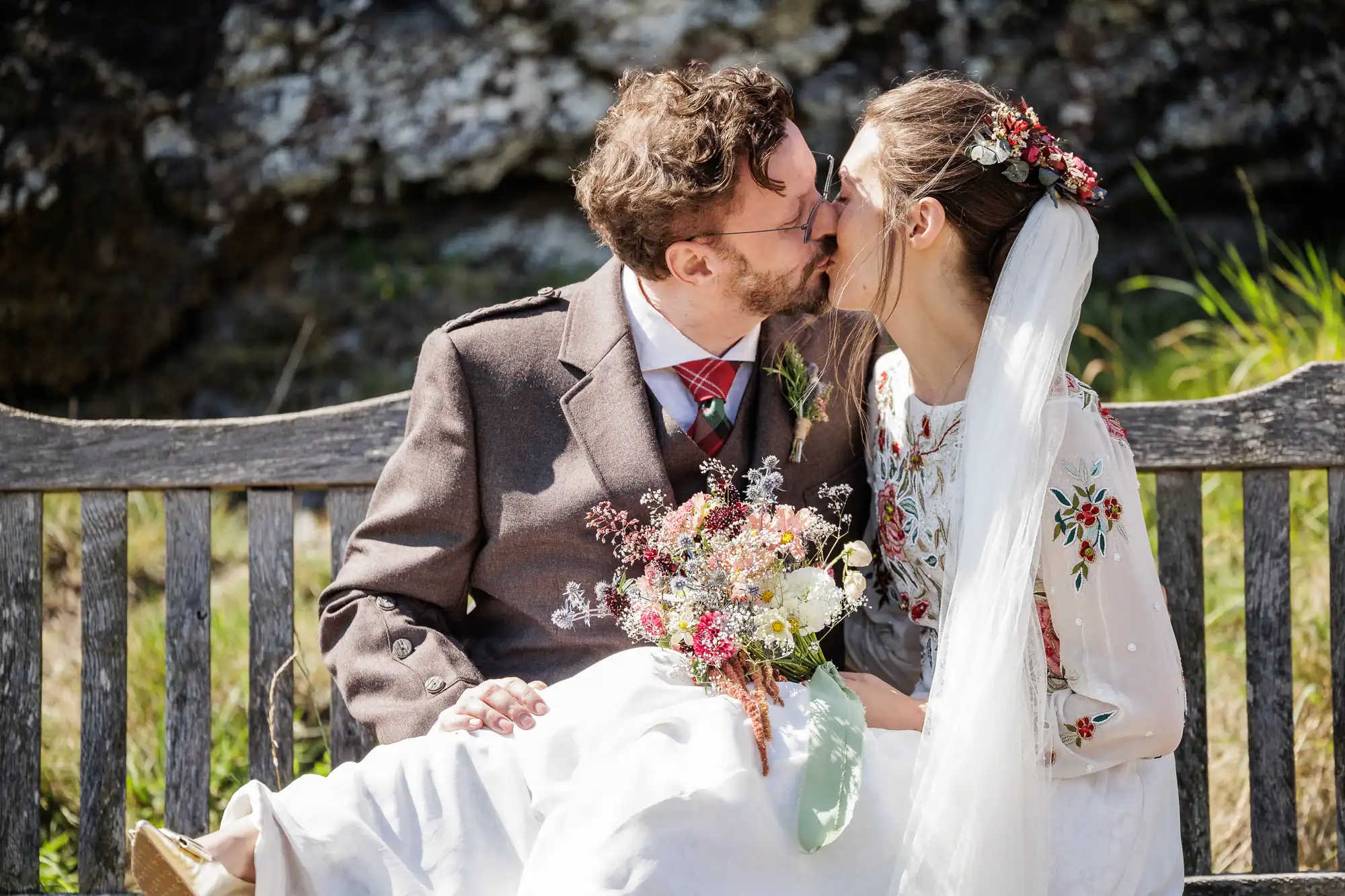 A couple in wedding attire share a kiss while sitting on a wooden bench outdoors. The bride wears a white dress with floral embroidery and a veil adorned with flowers. The groom wears a brown suit and a red tartan tie. The bride holds a colourful bouquet.