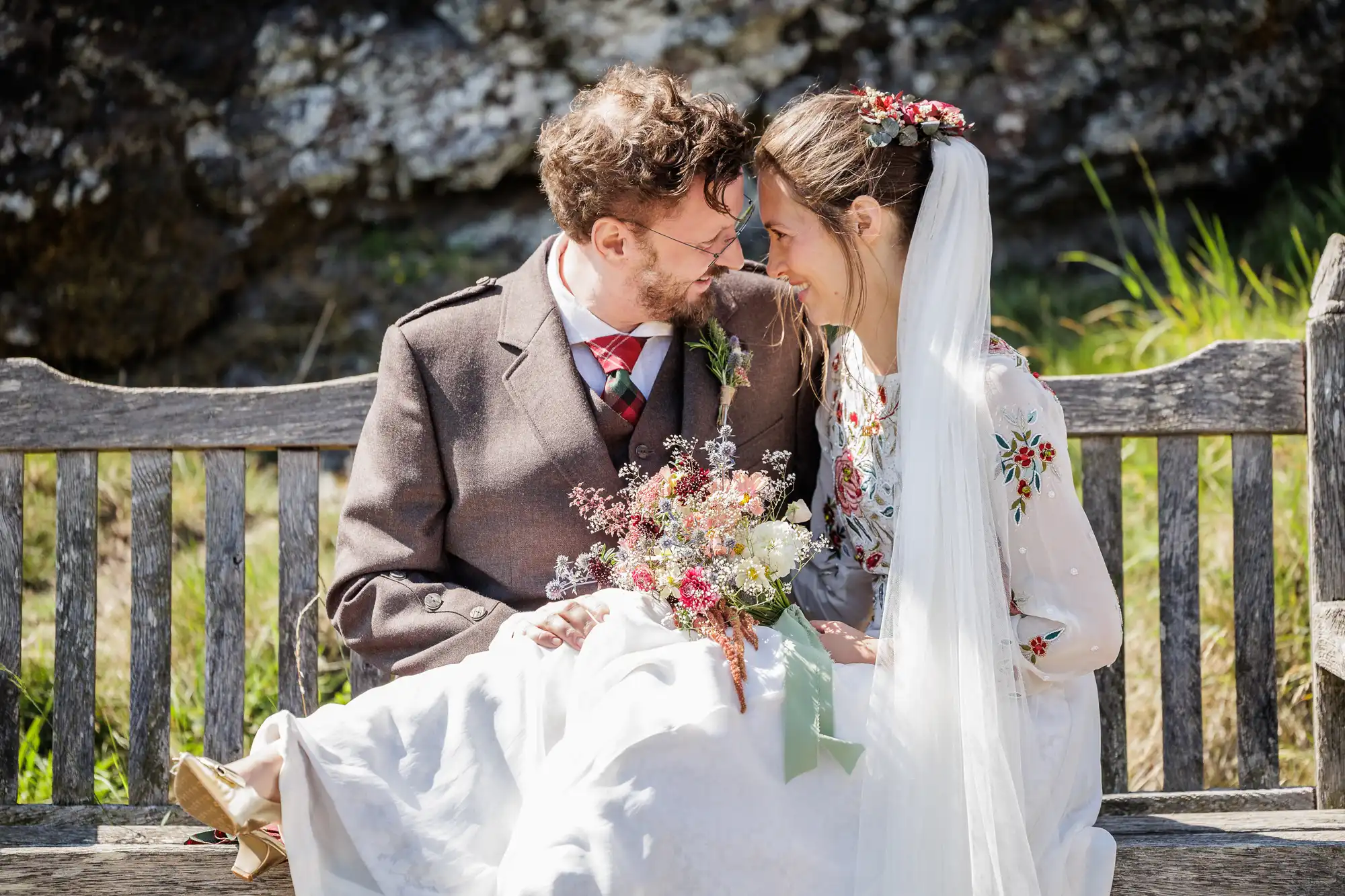 A bride and groom sit close on a wooden bench outdoors, touching foreheads and smiling. The bride wears a white floral dress, veil, and holds a bouquet of wildflowers. The groom wears a brown suit with a tartan tie. Sunlight highlights their joyful expressions.