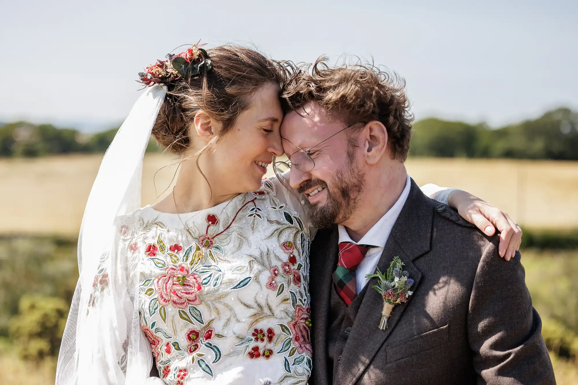 A smiling bride and groom stand outside in a sunlit field. The bride wears a floral embroidered dress, veil, and flower hairpiece, while the groom wears glasses and a brown suit with a tartan tie. They touch foreheads affectionately, eyes closed.