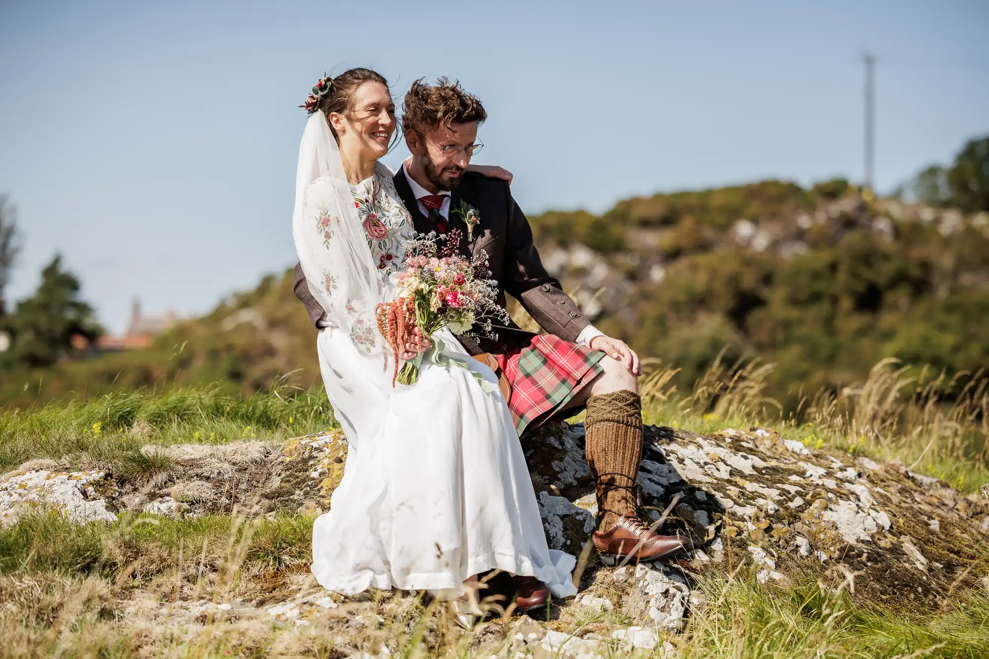 A smiling bride in a white dress and veil sits beside a groom in a dark jacket, red tartan kilt, and knee-high socks. They sit on a grassy rock outdoors in bright sunlight, with the bride holding a bunch of wildflowers.