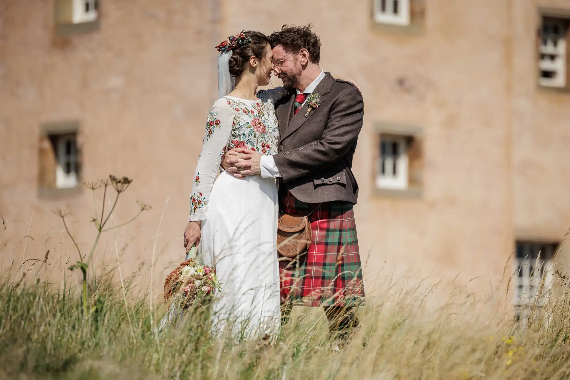 A bride and groom stand closely together outdoors, smiling and touching foreheads. The bride wears a white dress with colourful floral embroidery and holds a bouquet; the groom wears a brown jacket and a red-green tartan kilt. A stone building is in the background.