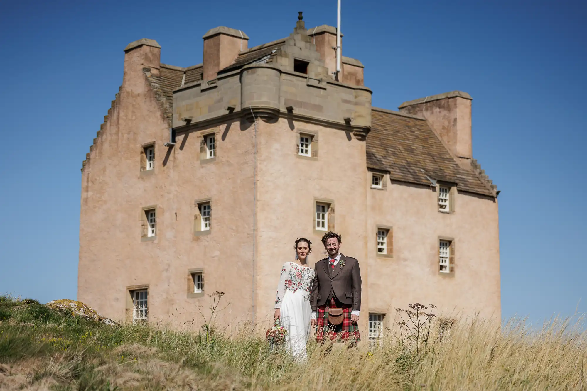A bride in a white dress and a groom in a brown jacket and red tartan kilt stand together in tall grass in front of a large, historic stone castle with small windows, under a clear blue sky.