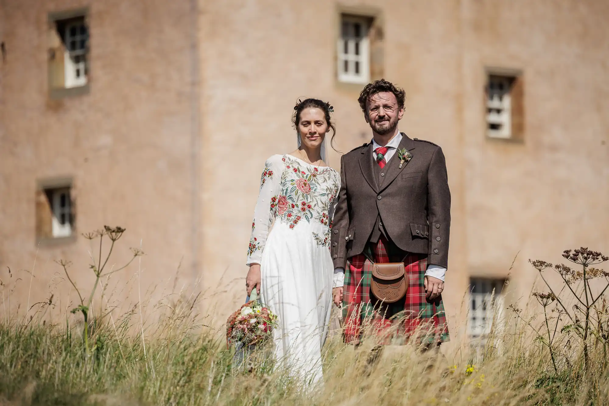 A man and woman stand in tall grass in front of a beige stone building. The woman wears a white, flower-embroidered dress and holds a bouquet. The man wears a brown jacket, red checked kilt, sporran, and tie. Both look at the camera, smiling slightly.