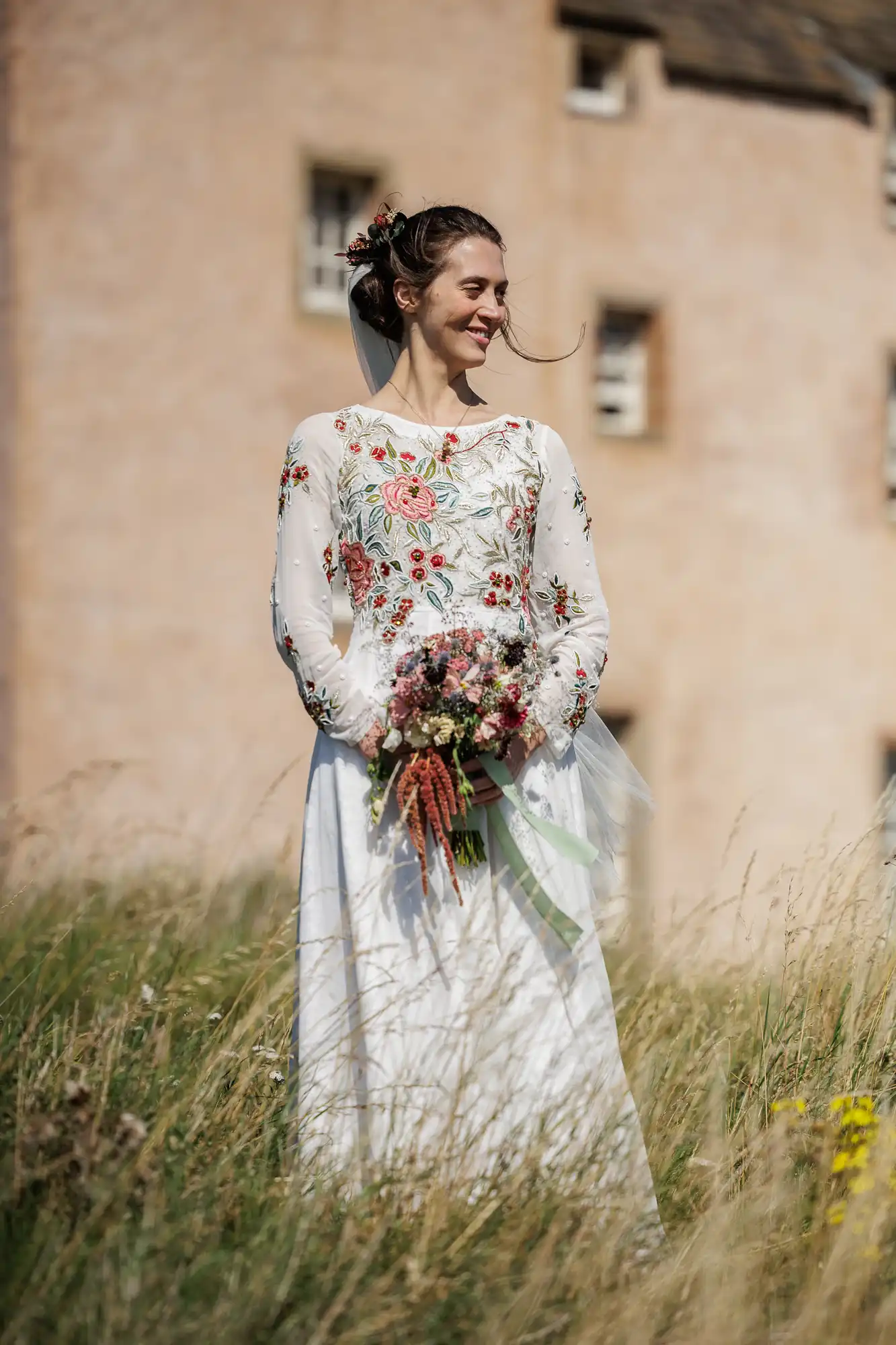 A smiling bride stands in tall grass, holding a bouquet. She wears a white dress with colourful floral embroidery, and her hair is styled with flowers. A rustic, light-brown building with small windows is blurred in the background under a bright sky.