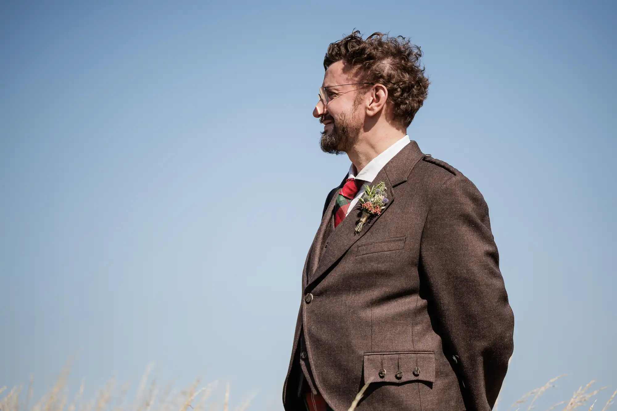 A man with curly hair and a beard, wearing glasses and a brown suit with a red tie and buttonhole, stands outdoors against a clear blue sky, looking to his left. Pale grasses are visible at the bottom of the image.