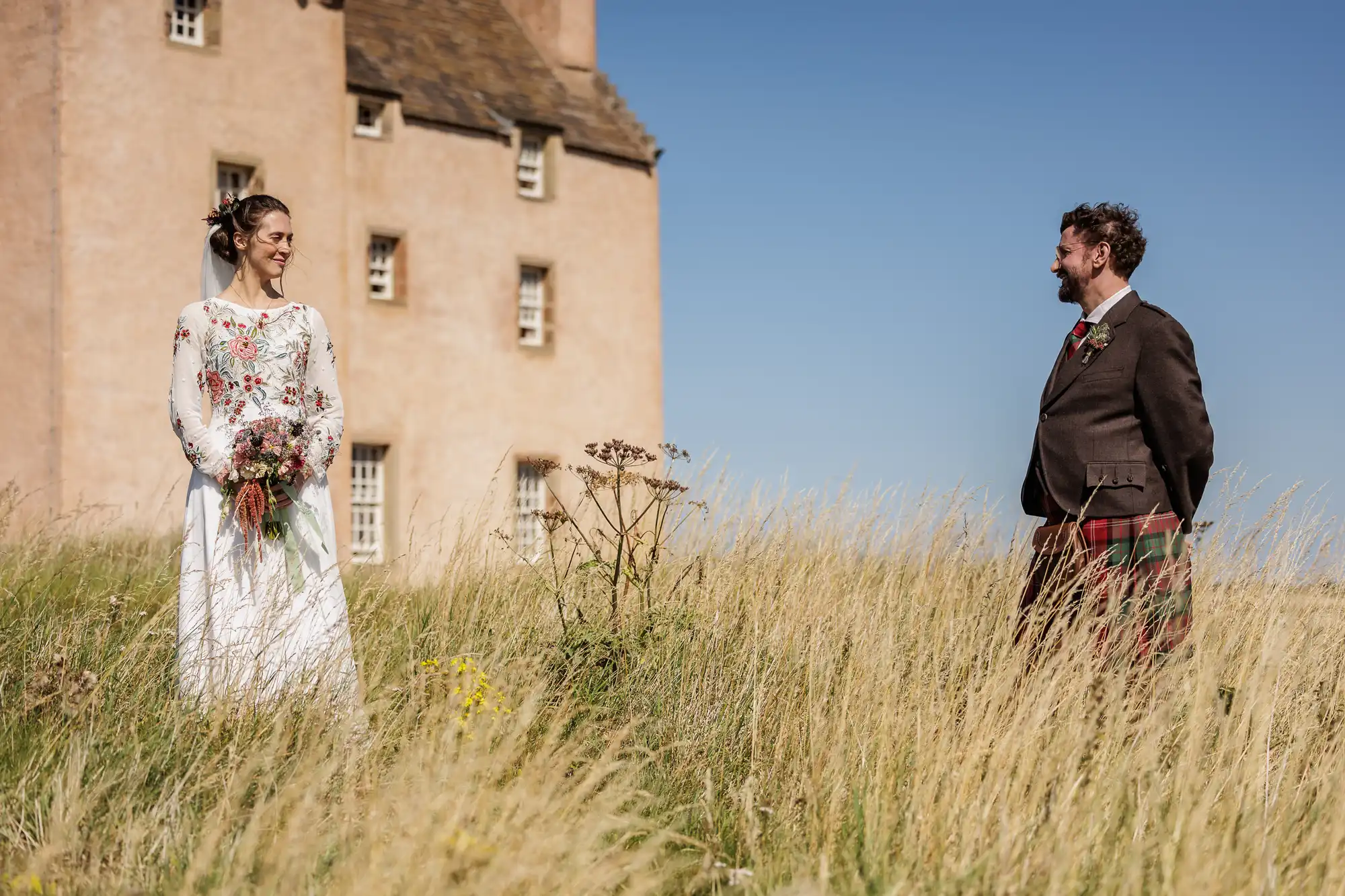 A smiling bride in a white floral dress holds a bouquet and faces a groom in a brown jacket and tartan kilt. They stand in tall grass, with a historic, pale stone building and clear blue sky in the background.