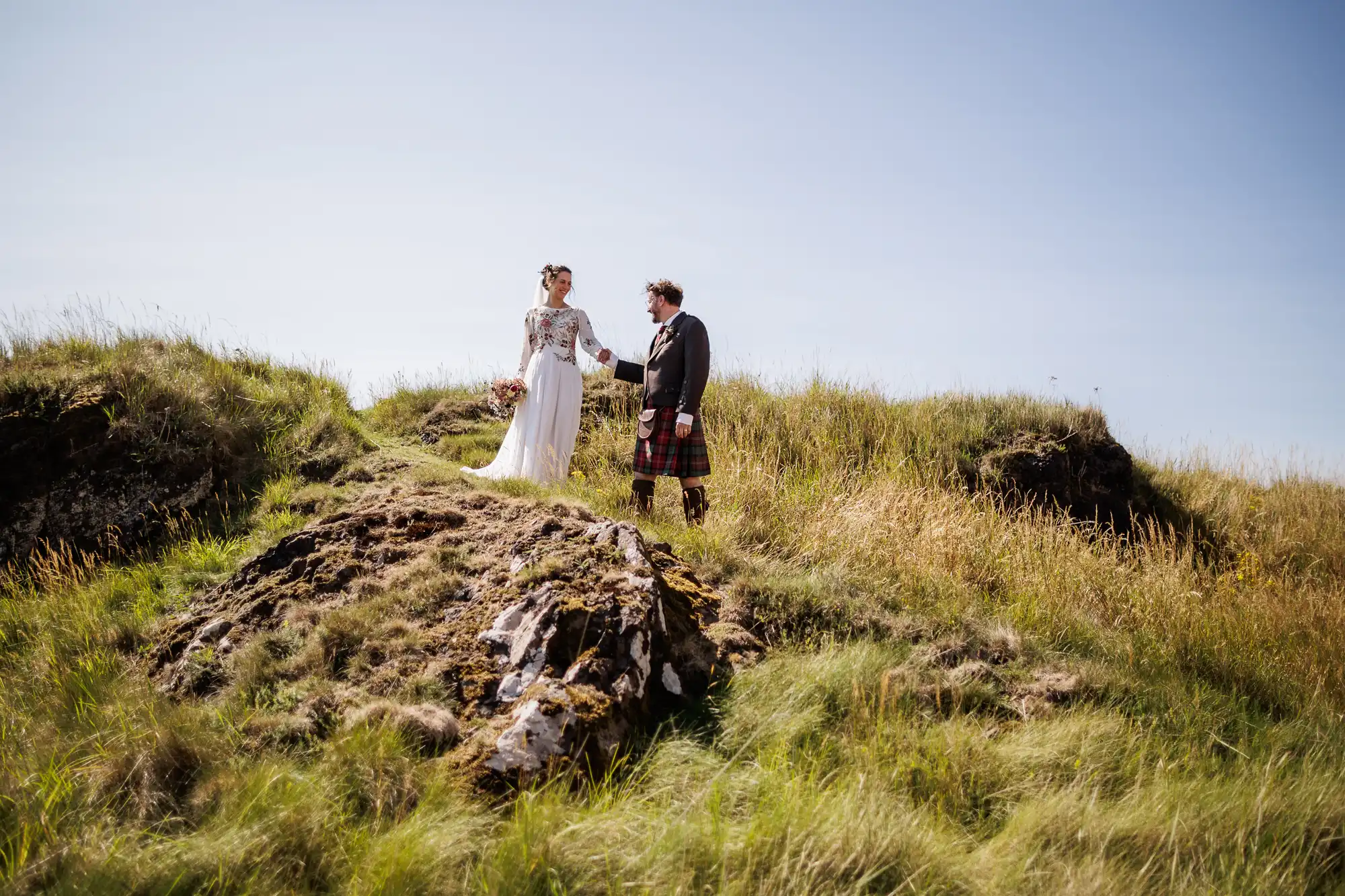 A bride in a white dress and a groom in a traditional Scottish kilt stand hand in hand on a grassy hill under a clear blue sky, surrounded by green grass and rugged rocks, creating a romantic outdoor scene.