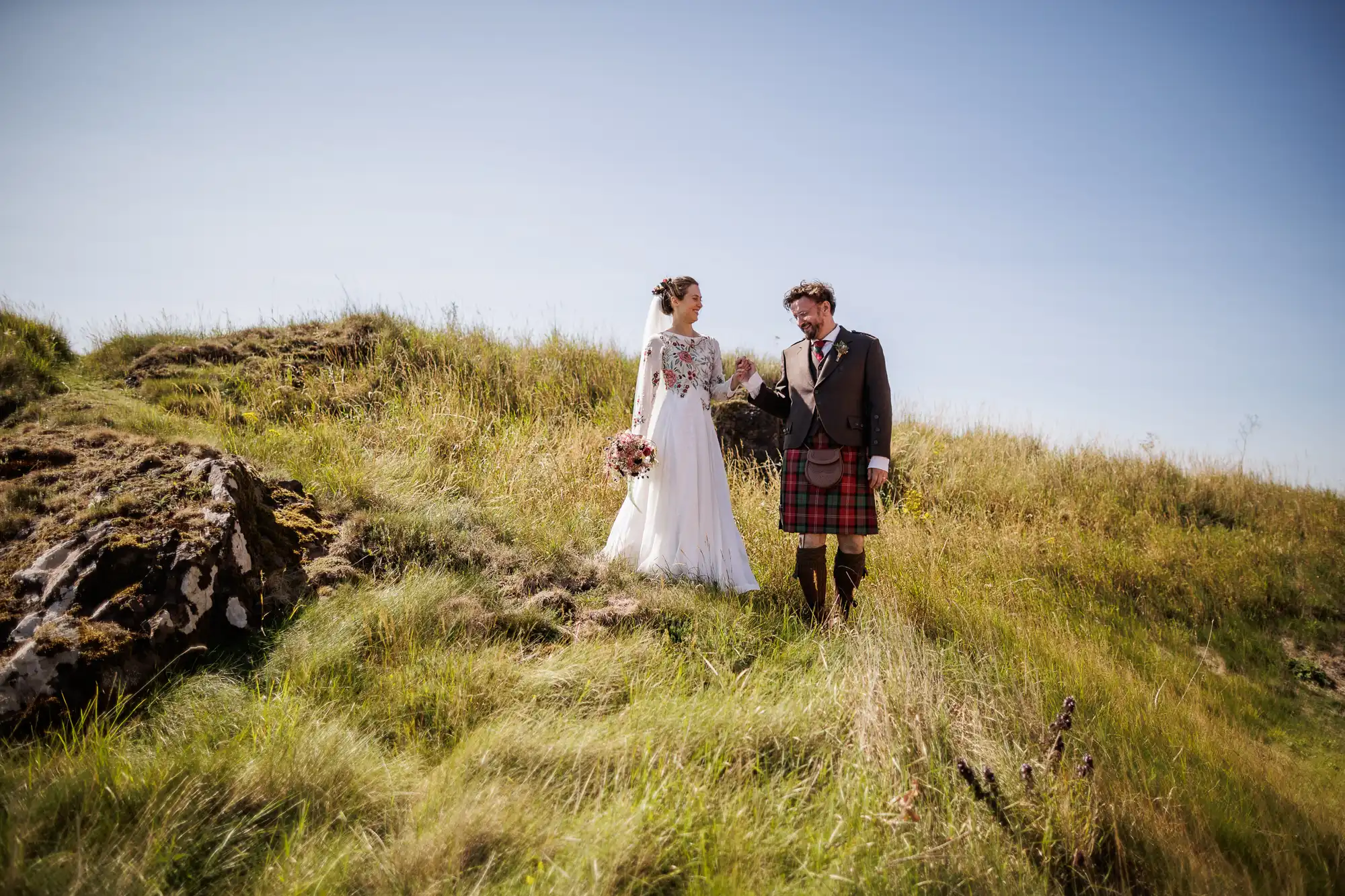 A bride in a white dress and a groom in a kilt stand on grassy, sunlit hills, holding hands and smiling at each other under a clear blue sky. The bride holds a bouquet, and wild grass surrounds them, creating a natural, romantic scene.