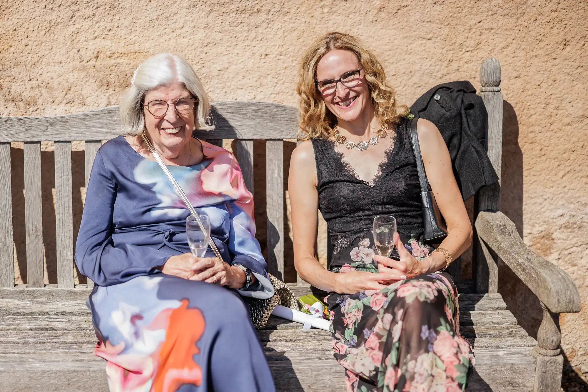 Two women sit smiling on a wooden bench outdoors. The older woman wears glasses and a long floral dress, holding a drink. The younger woman has curly blonde hair, a black sleeveless top, and a floral skirt, also holding a drink. Sunlight brightens the scene.