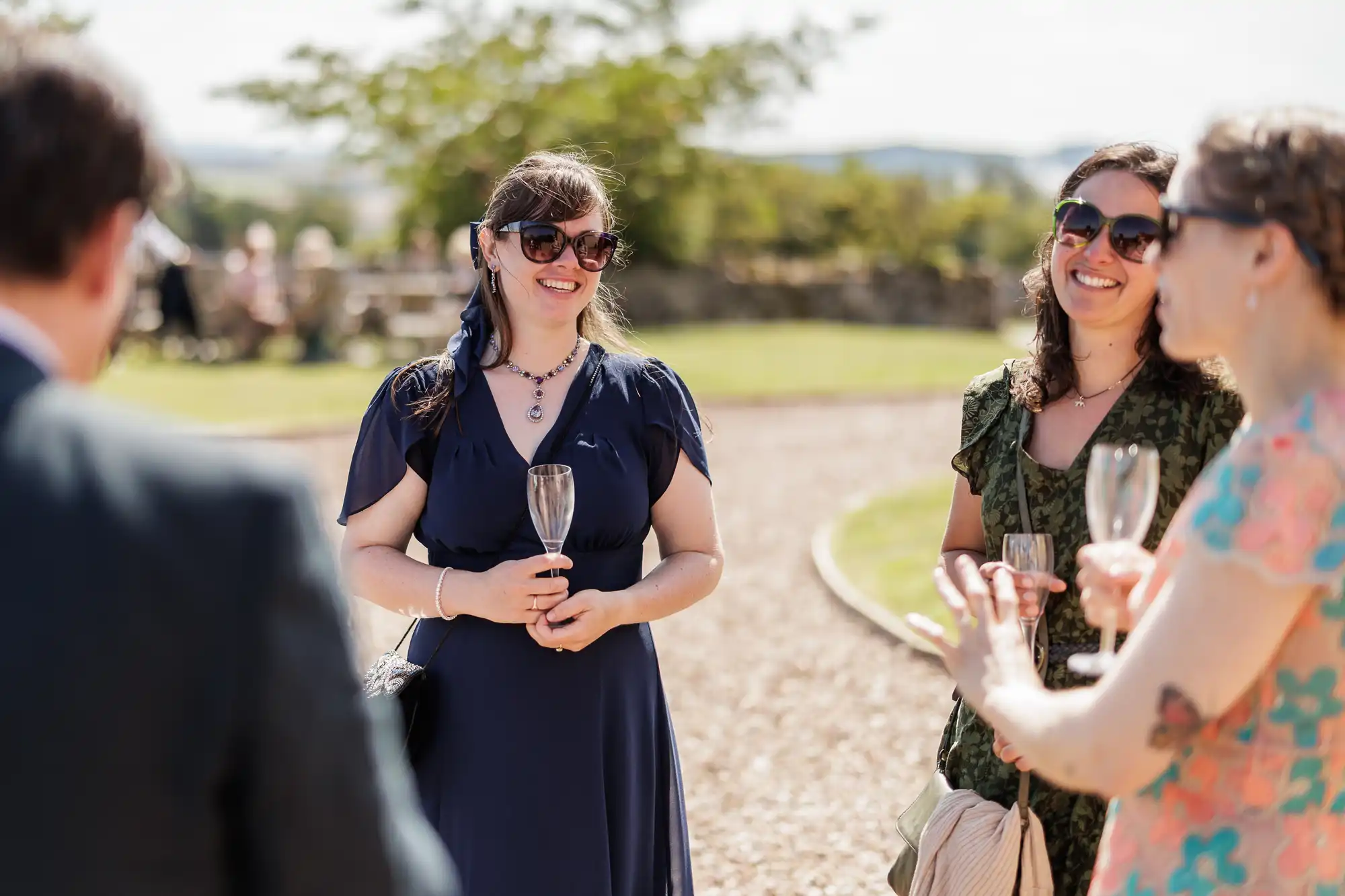 Four people standing outdoors on a sunny day, dressed in formal attire. Two women in sunglasses hold empty champagne glasses and smile. A third woman in a floral dress is seen from the side. The group is engaged in conversation, with trees in the background.