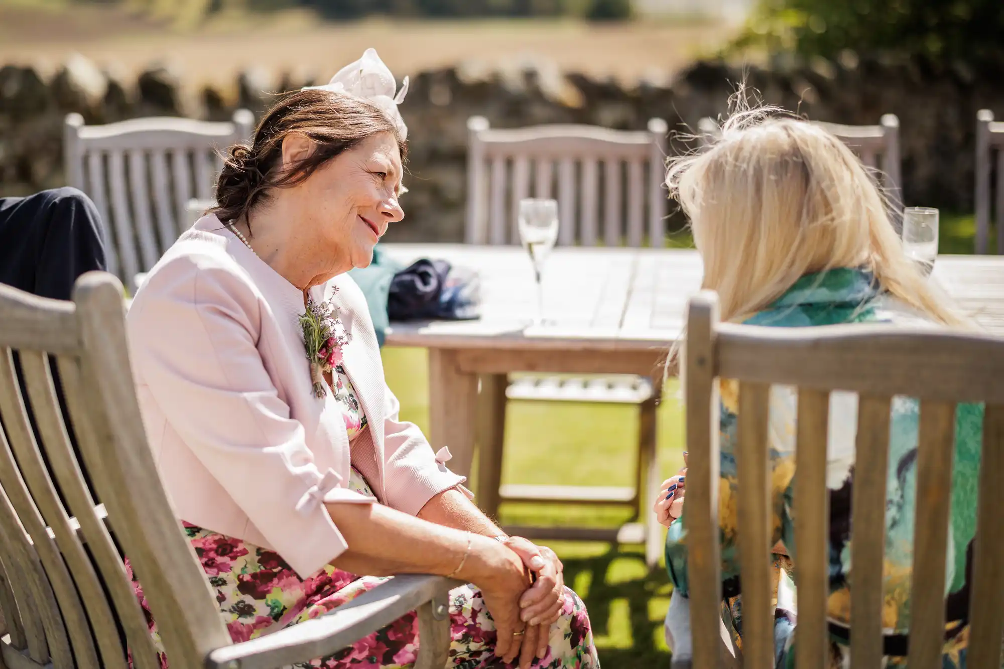 An older woman in a floral dress and pale pink jacket smiles warmly at a blonde woman seated next to her at an outdoor wooden table, with sunlit grass, stone wall, and empty champagne glasses in the background.