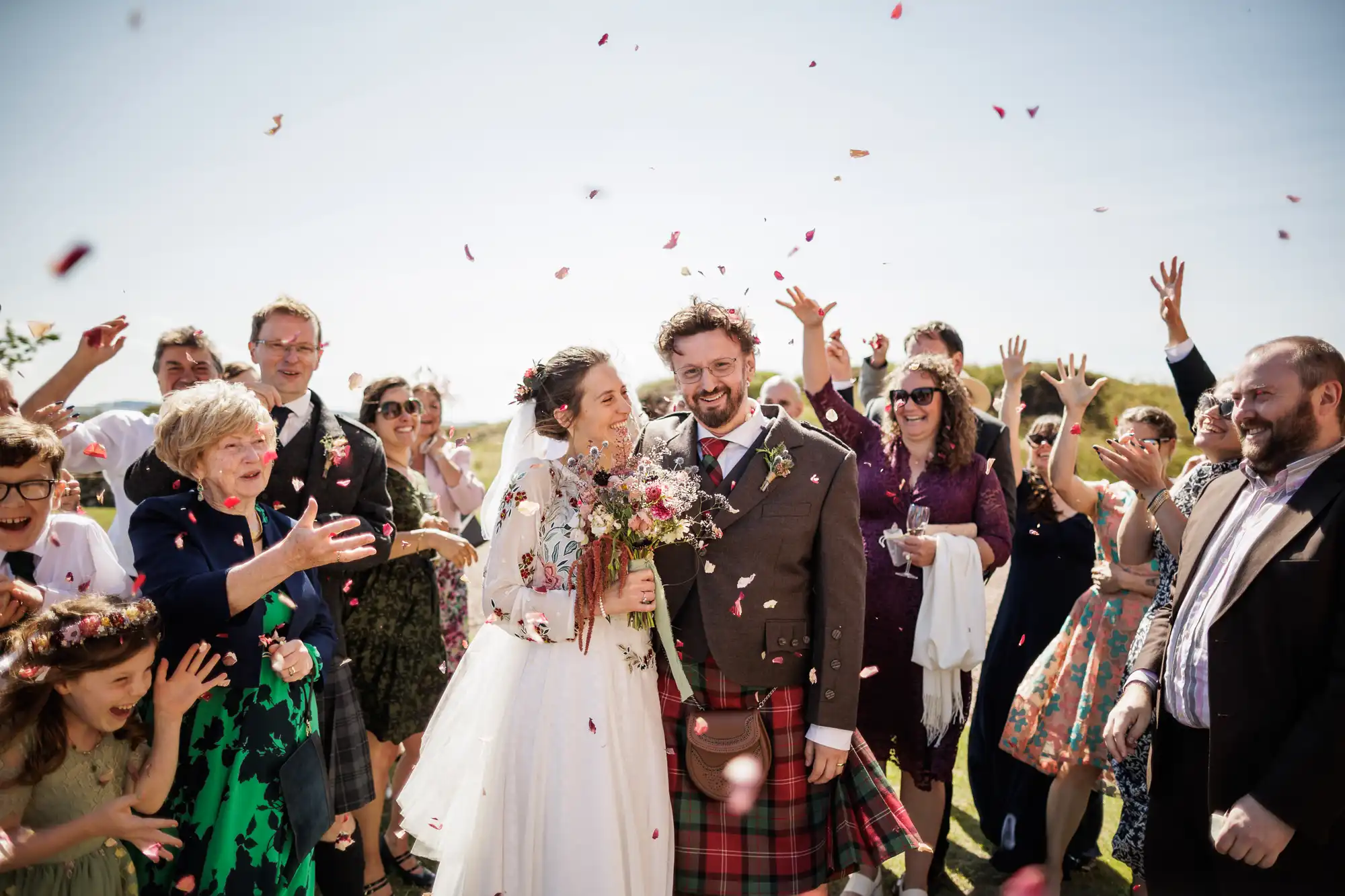A joyful bride and groom stand together outside, smiling as wedding guests throw flower petals. The bride holds a bouquet and wears a white dress; the groom wears a jacket and red tartan kilt. Friends and family cheer and celebrate around them under a sunny sky.