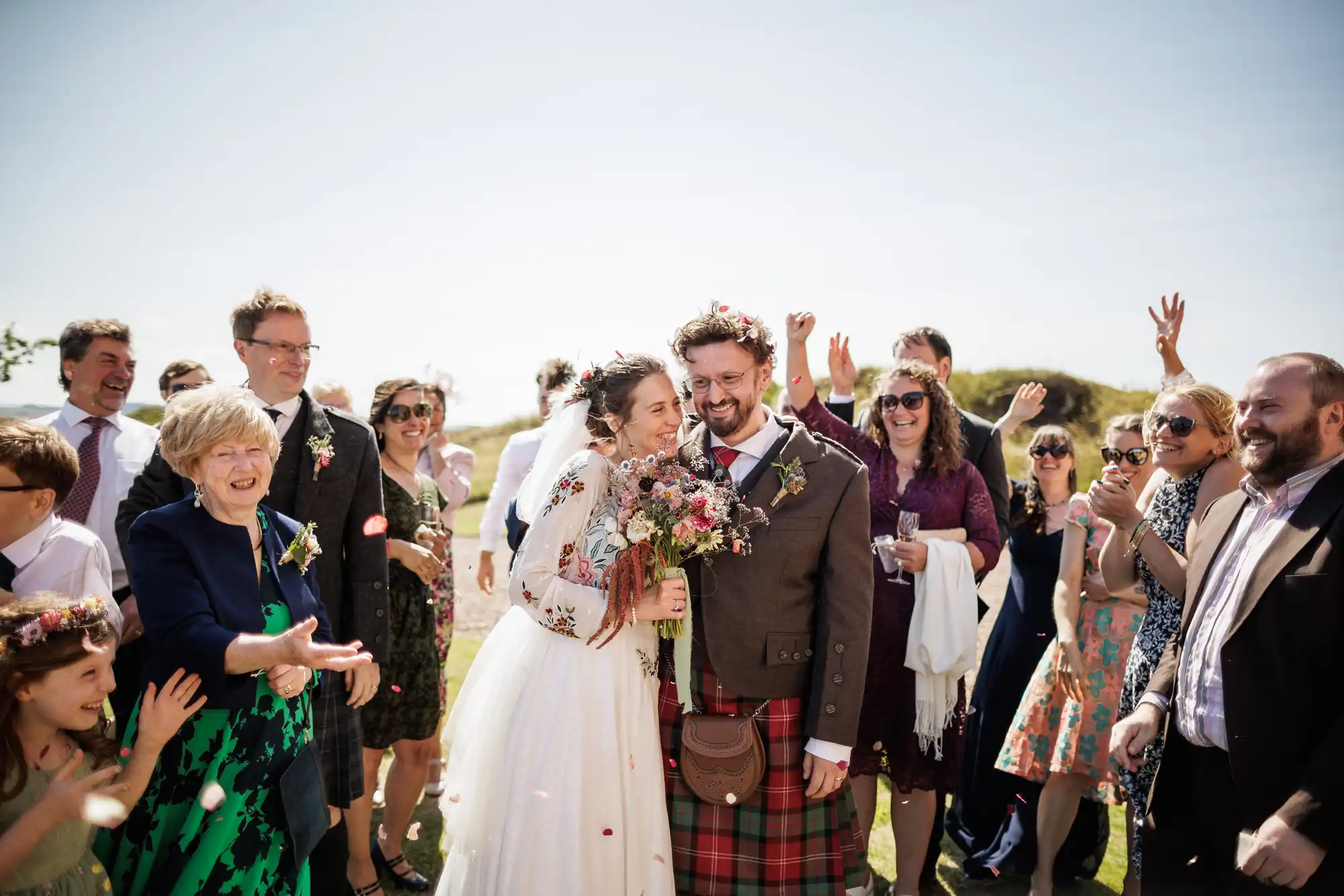 A joyful bride and groom stand together outside, surrounded by smiling guests. The bride wears a white dress and holds a bouquet; the groom wears a tartan kilt and brown jacket. Guests throw confetti and cheer under a clear, sunny sky.