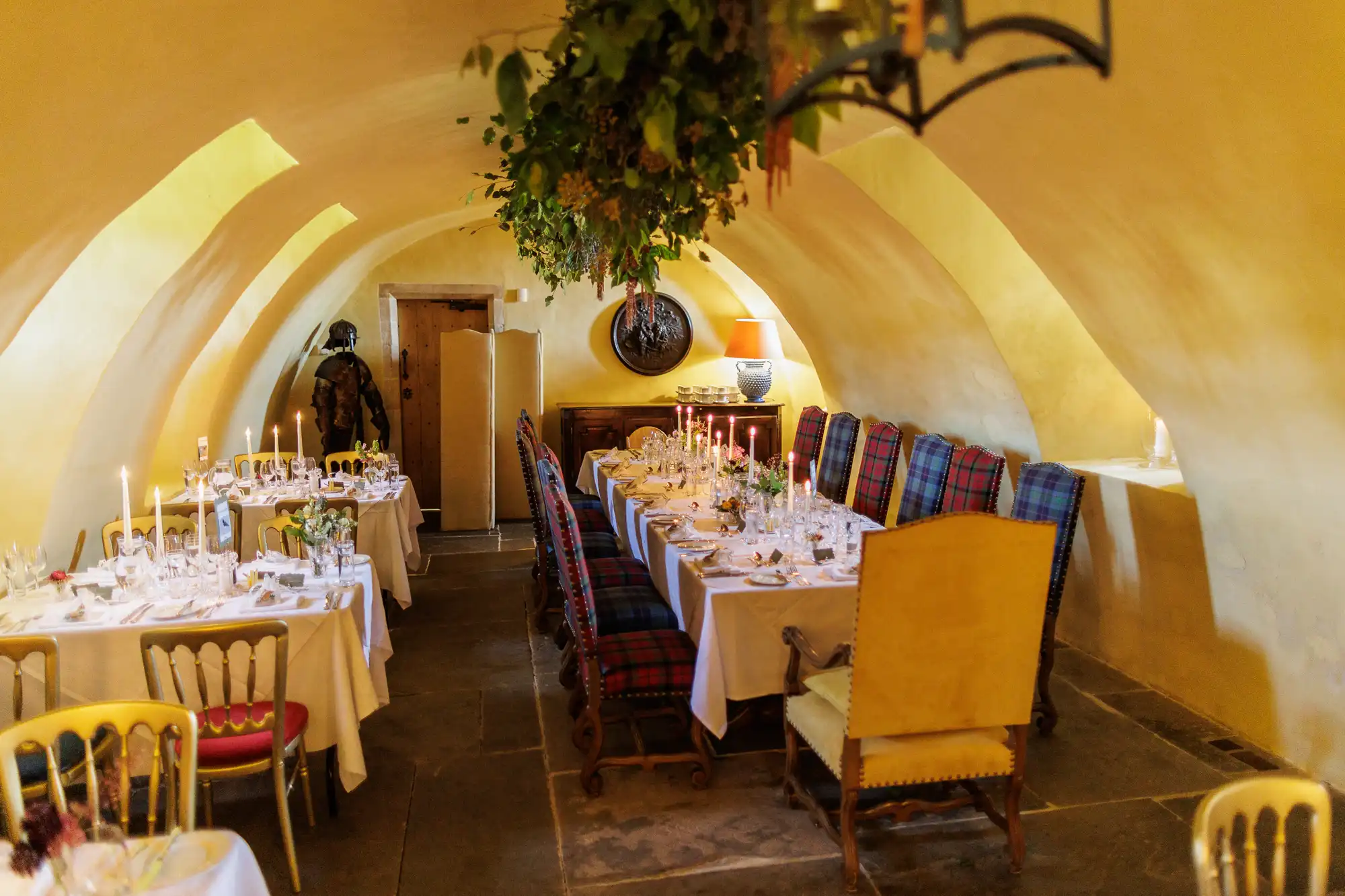 A warmly lit dining room with arched yellow walls, long tables set with white cloths, elegant glassware, and candles. The chairs are upholstered in red and blue check. Plants hang from the ceiling, and a person stands near a wooden door at the back.