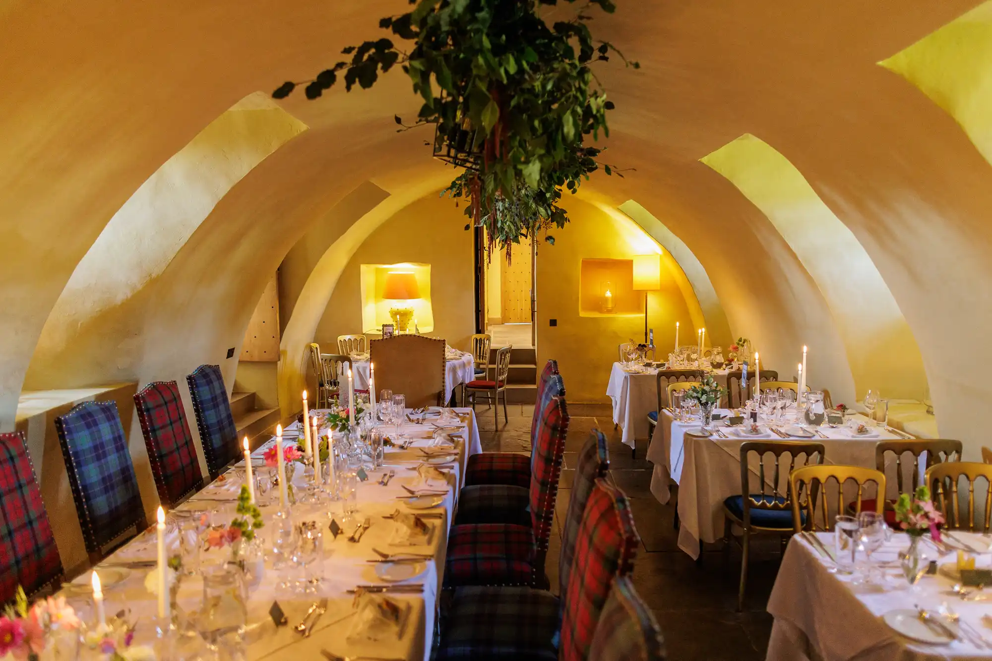 Elegant dining room with arched ceilings, checked chairs, and tables laid with white linen, glassware, and candles. Warm, ambient lighting and greenery hanging from the ceiling create a cosy, inviting atmosphere. Flowers decorate the tables.