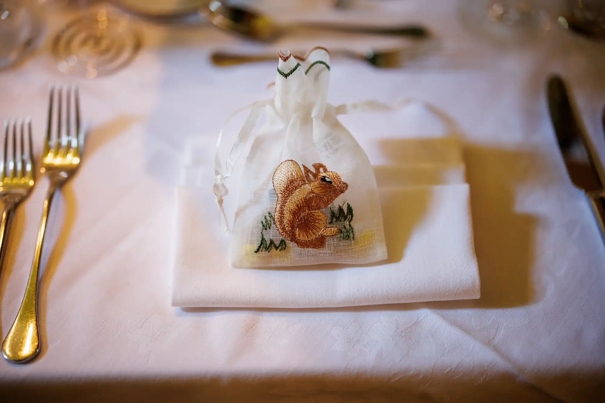 A small white drawstring bag with an embroidered squirrel sits on a folded white serviette, arranged on a formal dining table set with cutlery and glassware.