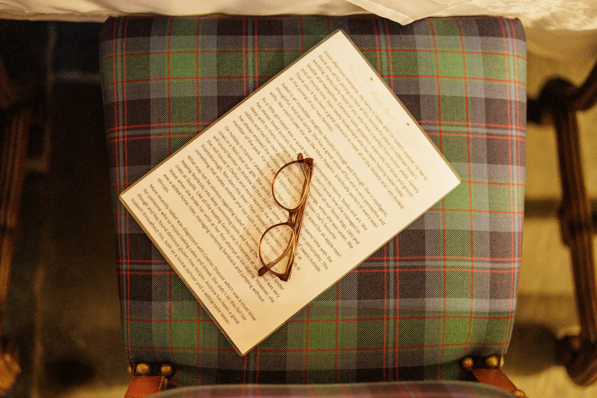 A pair of brown spectacles rests on a printed page full of text, placed diagonally on a blue, green, and red checked-patterned upholstered chair. The image is viewed from above, showing the entire seat and partial legs of the chair.