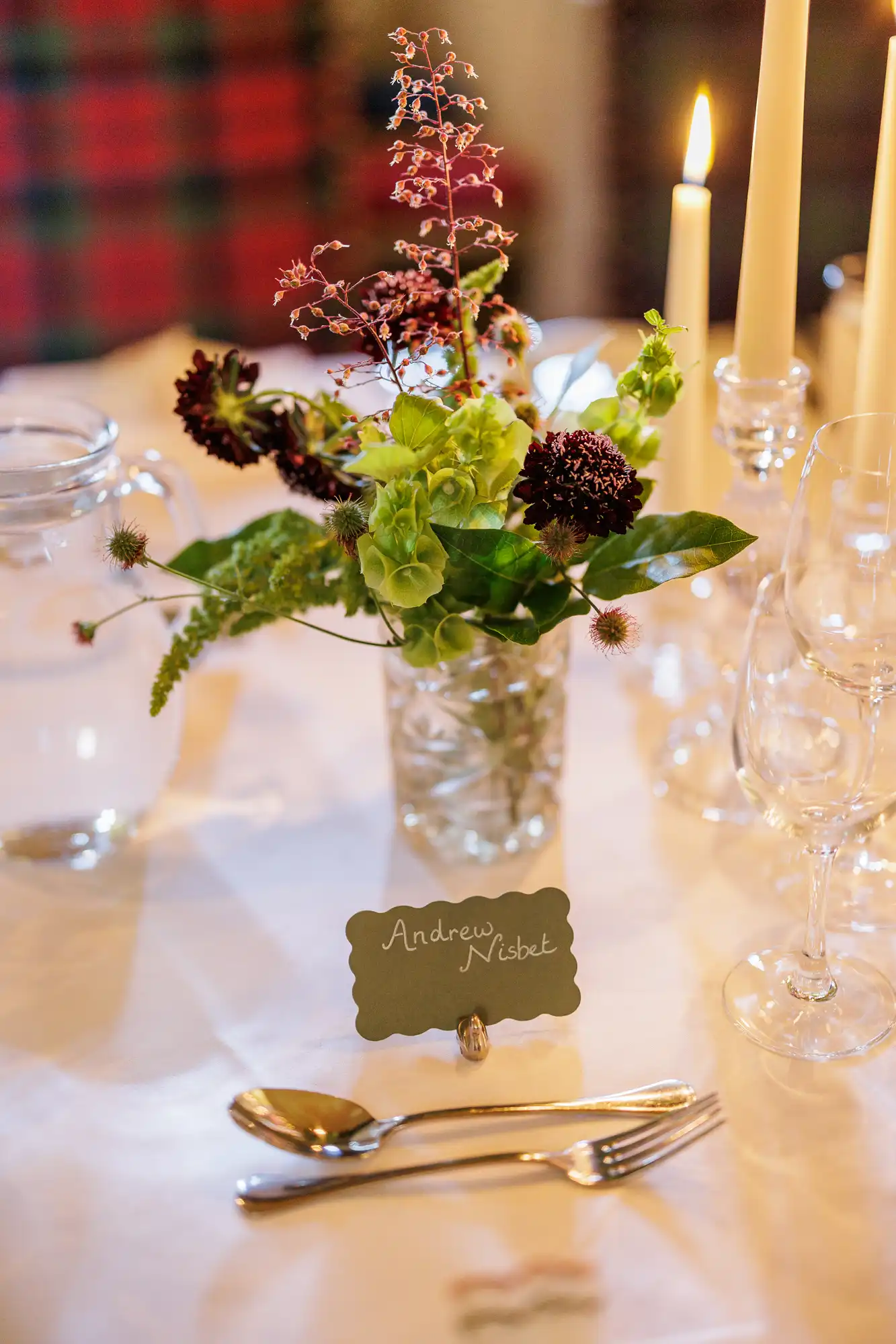 A formal table setting with a small bouquet of green and purple flowers in a glass vase, a lit taper candle, clear glassware, silver cutlery, and a name card reading "Andrew Nisbet" placed at the centre. The scene is softly lit and elegant.