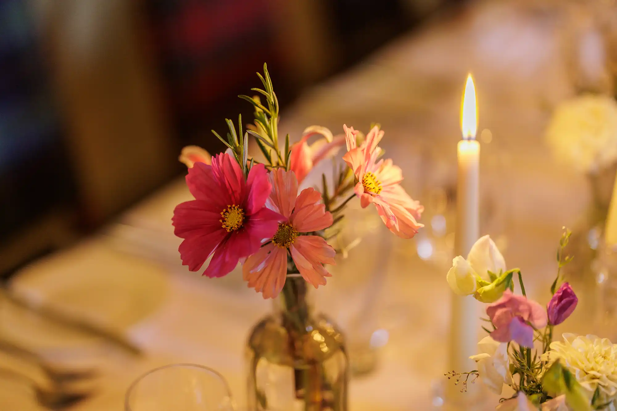 A small glass vase holds pink cosmos flowers and greenery on a softly lit table set for dining. Nearby, a white taper candle burns, surrounded by blurred glassware and cutlery, creating a warm, intimate atmosphere.