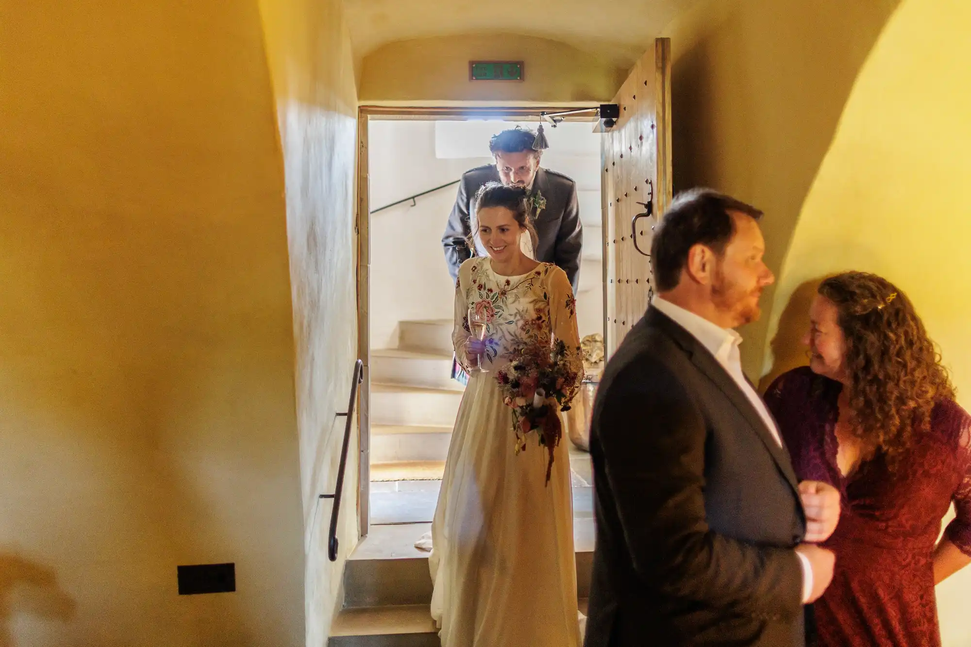 A bride in a long white dress with floral patterns enters a warmly lit room, smiling and holding a bouquet. A man in a suit follows her. In the foreground, a man and woman, both dressed formally, stand together and face each other, talking and smiling.