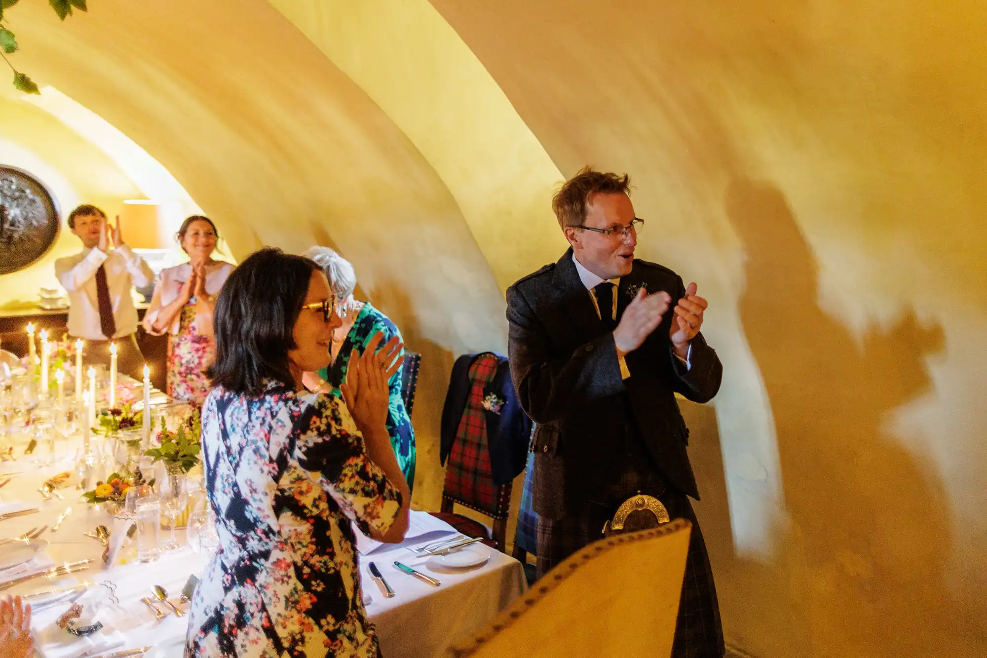 People stand and clap in a warmly lit room with yellow arched walls. A man in a suit and kilt smiles whilst applauding. Others, including a woman in a floral dress, are also applauding around a long table laid for a formal meal with candles and flowers.