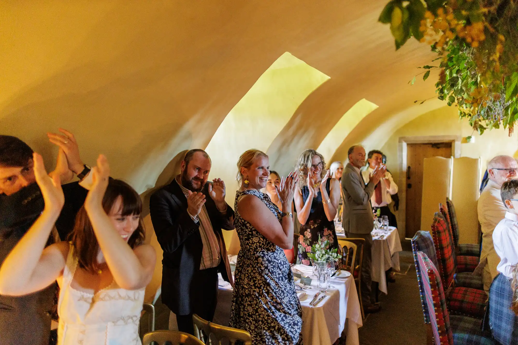 A group of well-dressed guests stand and applaud in a warmly lit, arched-ceilinged room decorated with flowers. Tables are set with white cloths and floral centrepieces, and everyone looks happy and engaged in the celebration.