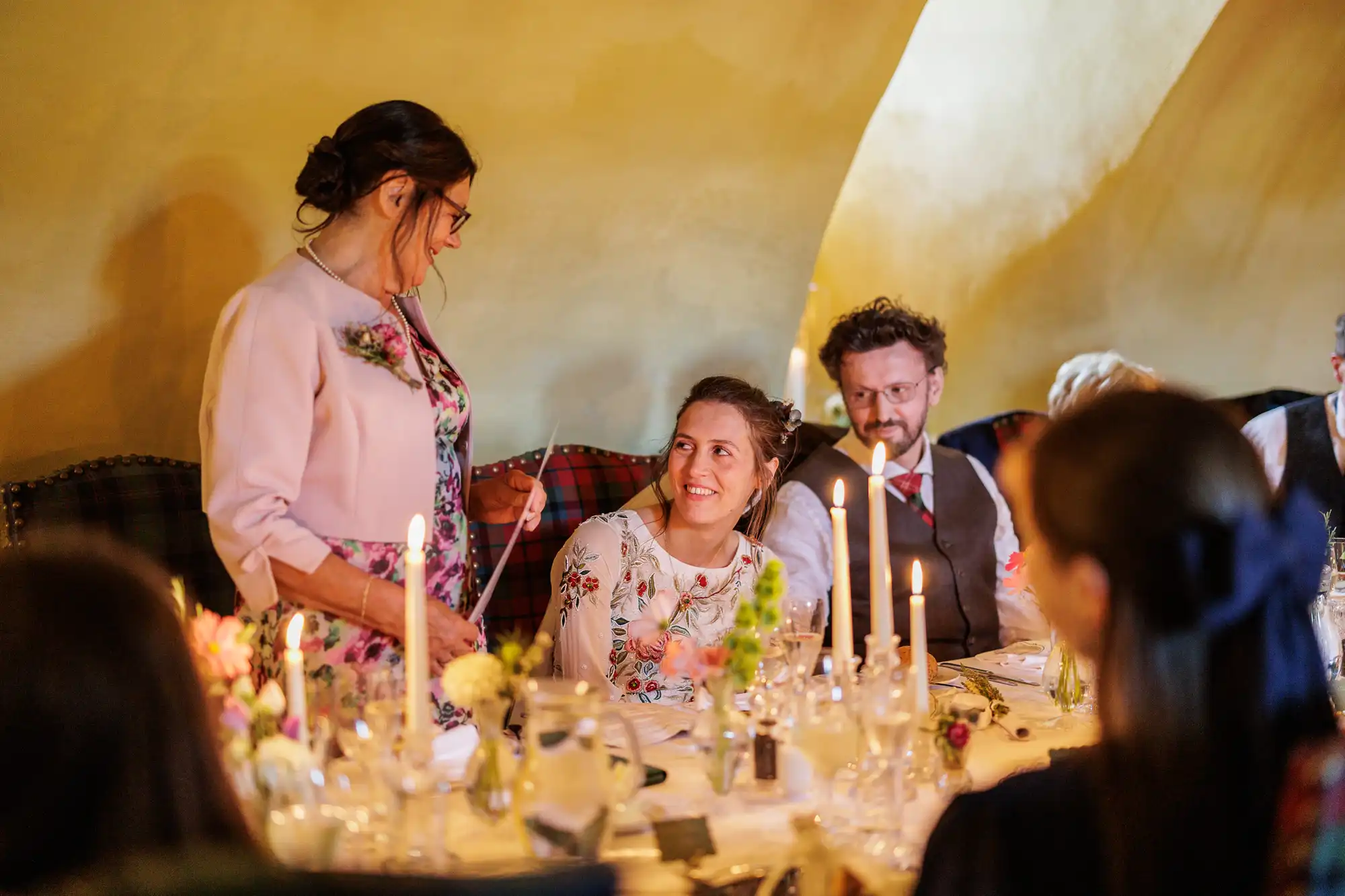 A woman stands giving a speech at a candlelit wedding reception table. She faces a smiling seated woman in a floral dress. Other guests watch, surrounded by flowers, candles, and glassware. The scene feels warm and celebratory, with soft lighting and a cosy ambience.