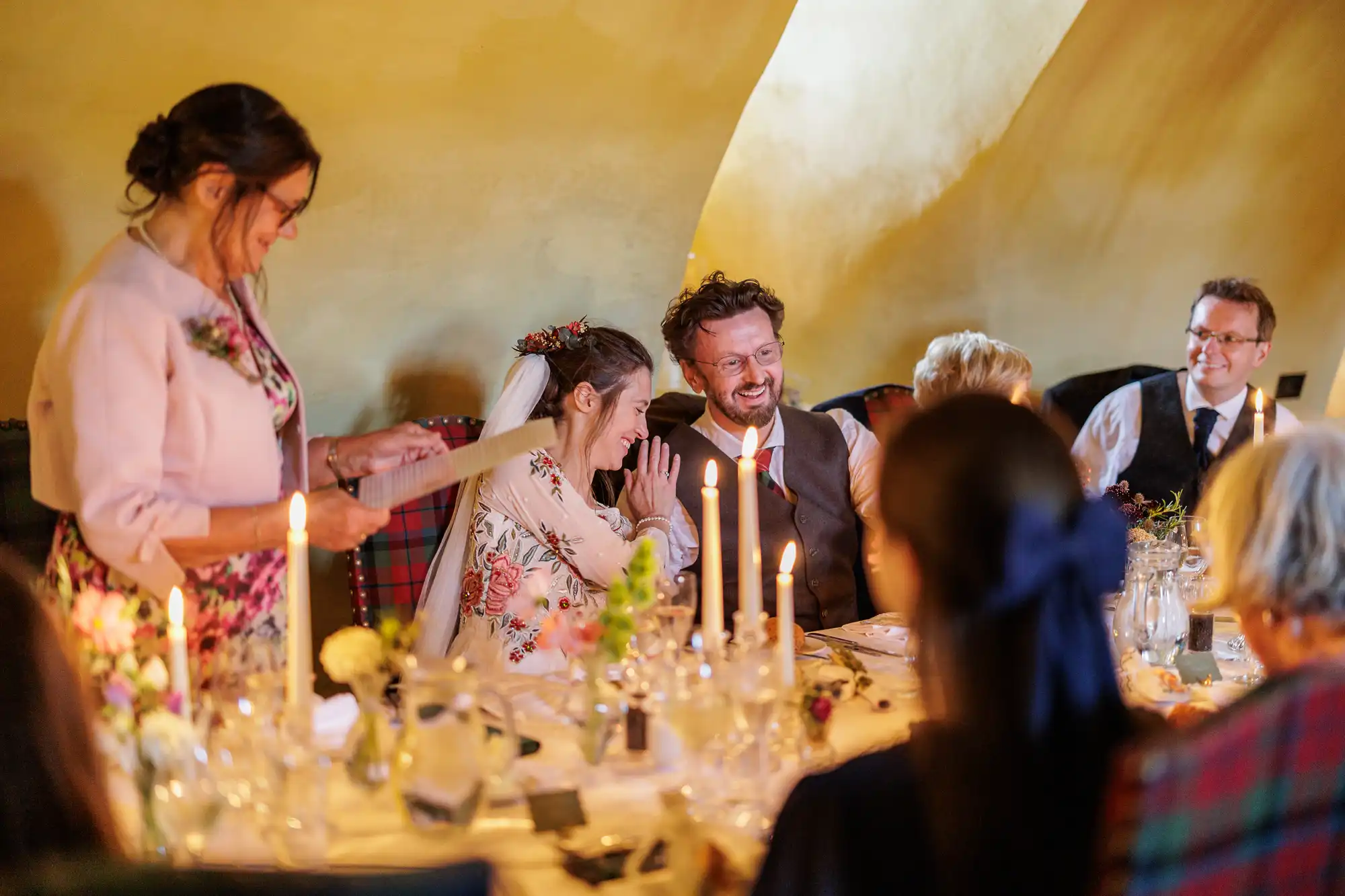 A smiling bride and groom sit at a candlelit table, surrounded by guests. The bride covers her mouth, possibly laughing or emotional, as a woman standing nearby reads from a sheet of paper. The room glows with warm, yellow light.