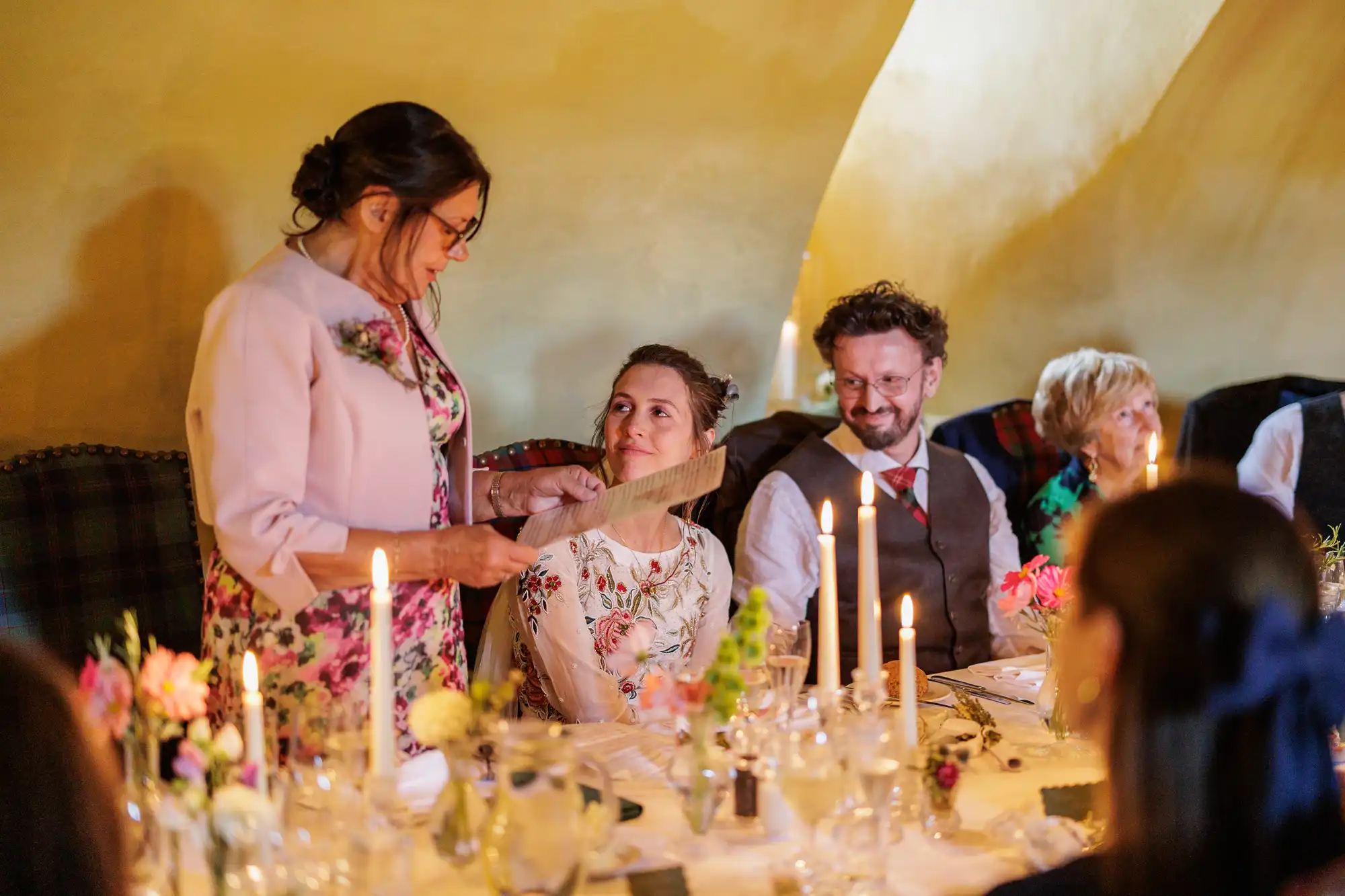 A woman stands reading from a piece of paper at a warmly lit dining table, while a bride in a floral dress smiles up at her. A man in a waistcoat and other guests sit nearby. The table is set with candles, flowers, and glassware, creating a festive atmosphere.