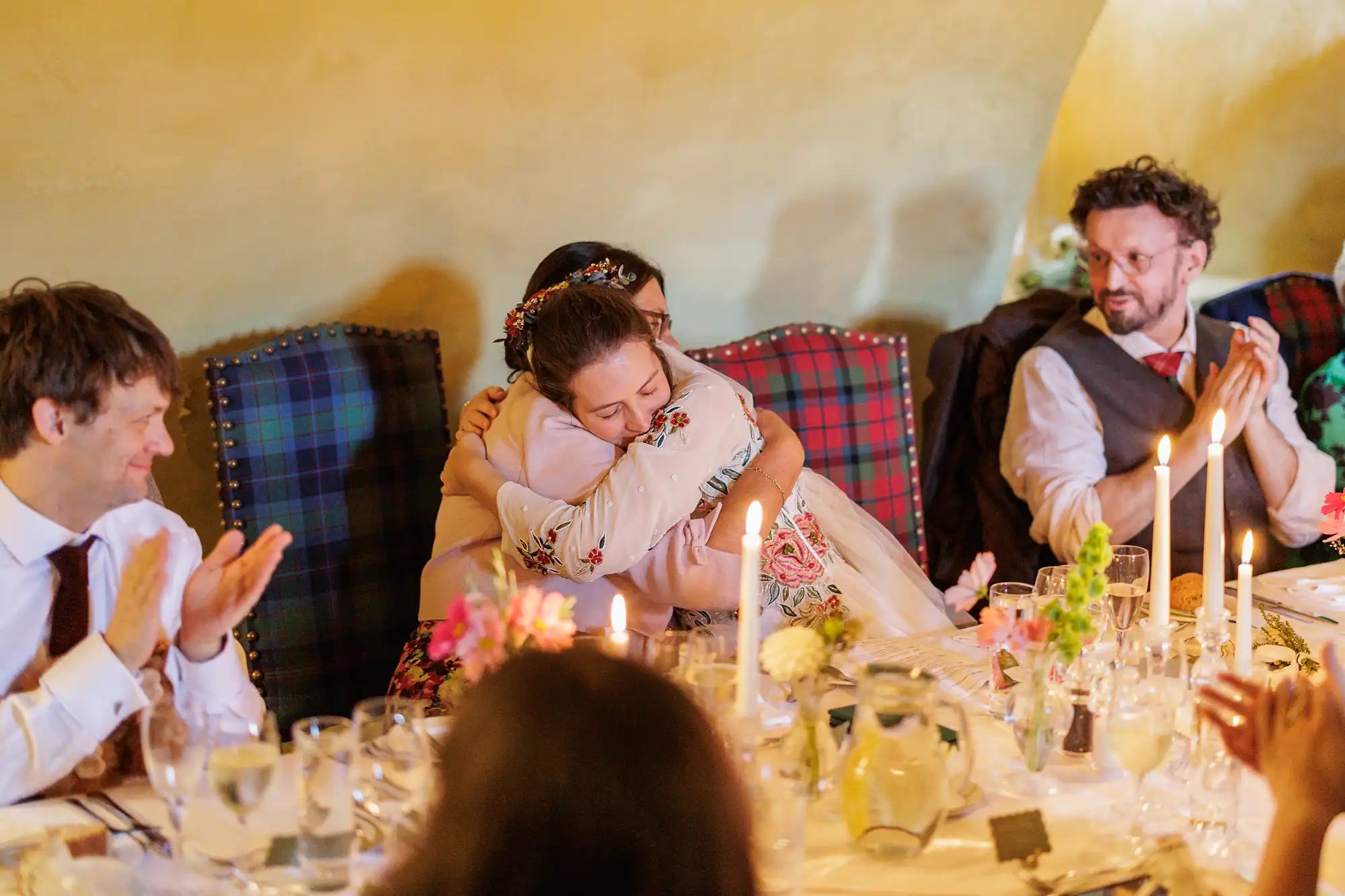 A bride warmly hugs a guest at a candlelit wedding reception table, while others seated nearby smile and clap. The scene features floral centrepieces, glasses, and tartan-upholstered chairs, creating a festive and intimate atmosphere.