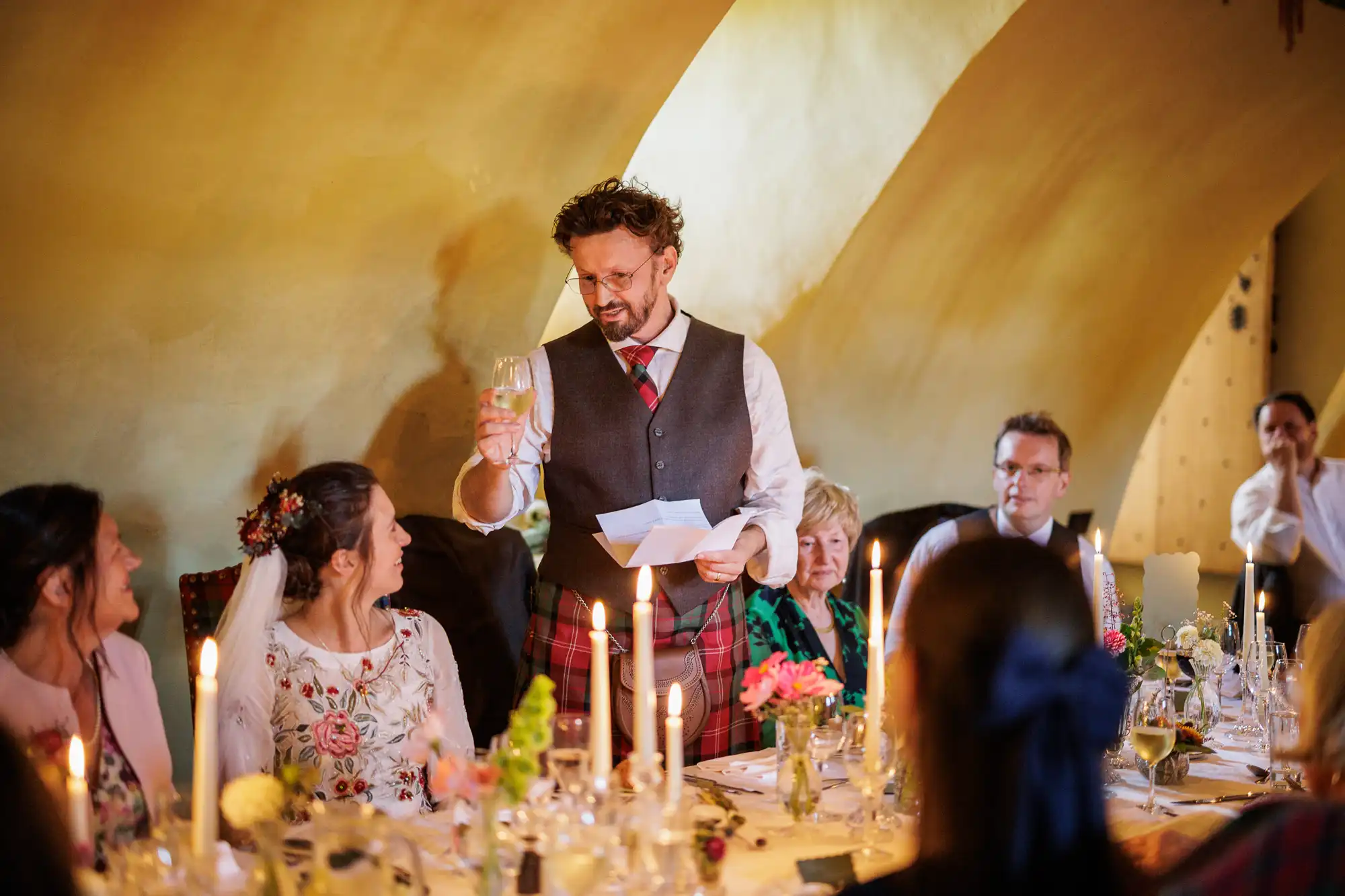 A man in a waistcoat and tartan kilt stands at a candlelit table, holding up a glass in a toast. He faces a seated woman in a white floral dress and veil. Guests smile around the decorated table, beneath warm, arched lighting, at what appears to be a wedding.