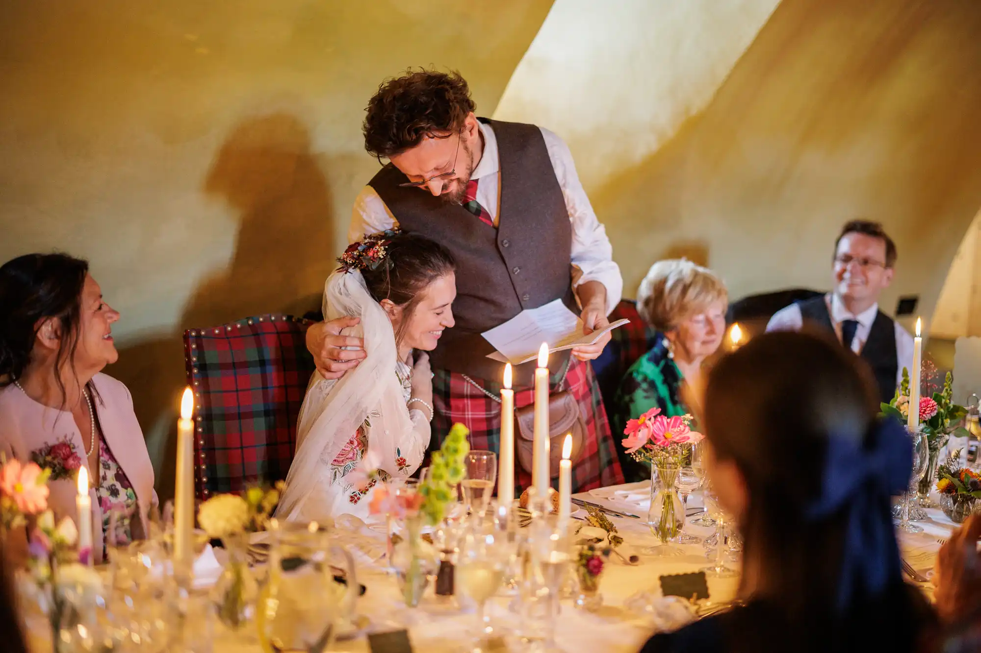 A man in a suit stands, hugging a seated woman in a wedding dress at a candlelit table. Guests around them smile warmly. The table is decorated with flowers, glasses, and candles, creating a festive, intimate atmosphere.