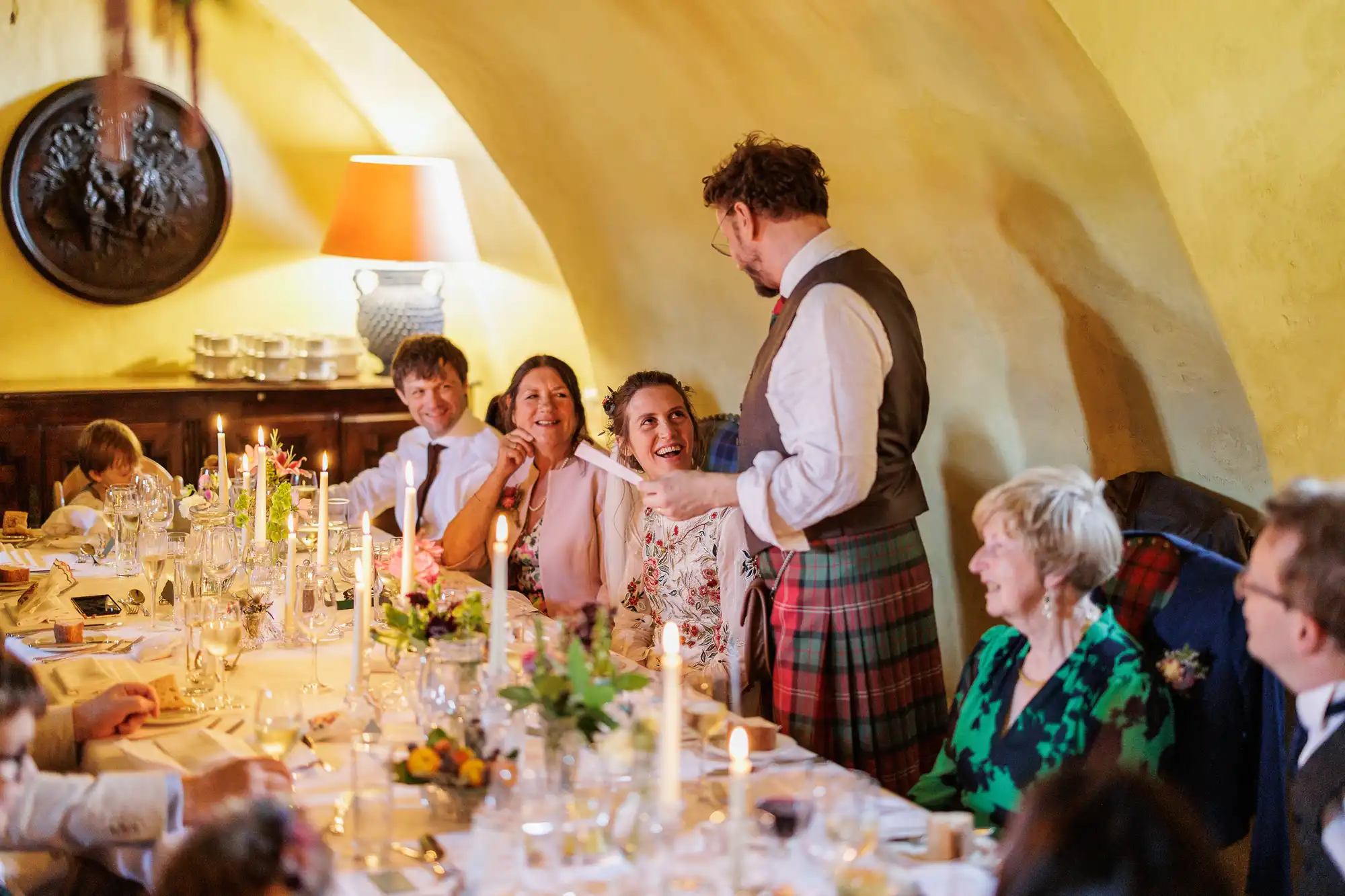 A man in a kilt stands and speaks to smiling guests seated at a long candlelit table set for a meal in a warmly lit room with yellow walls. Flowers, glasses, and plates decorate the table; everyone appears cheerful and engaged.