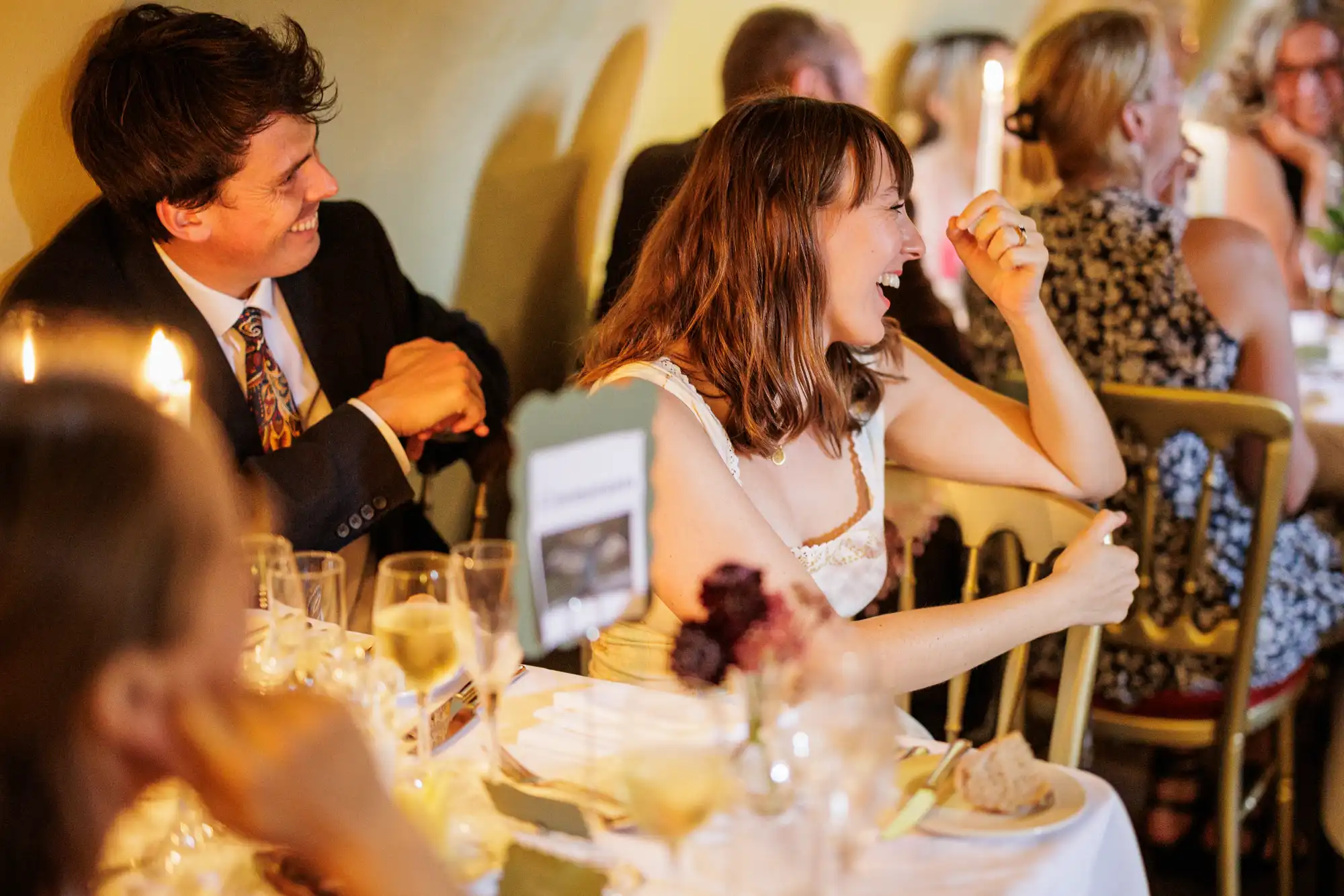 A woman in a white dress laughs joyfully at a table set for a formal dinner, whilst a man in a suit beside her smiles. Other guests seated at the table are blurred, with candles and glasses visible, creating a warm, celebratory atmosphere.