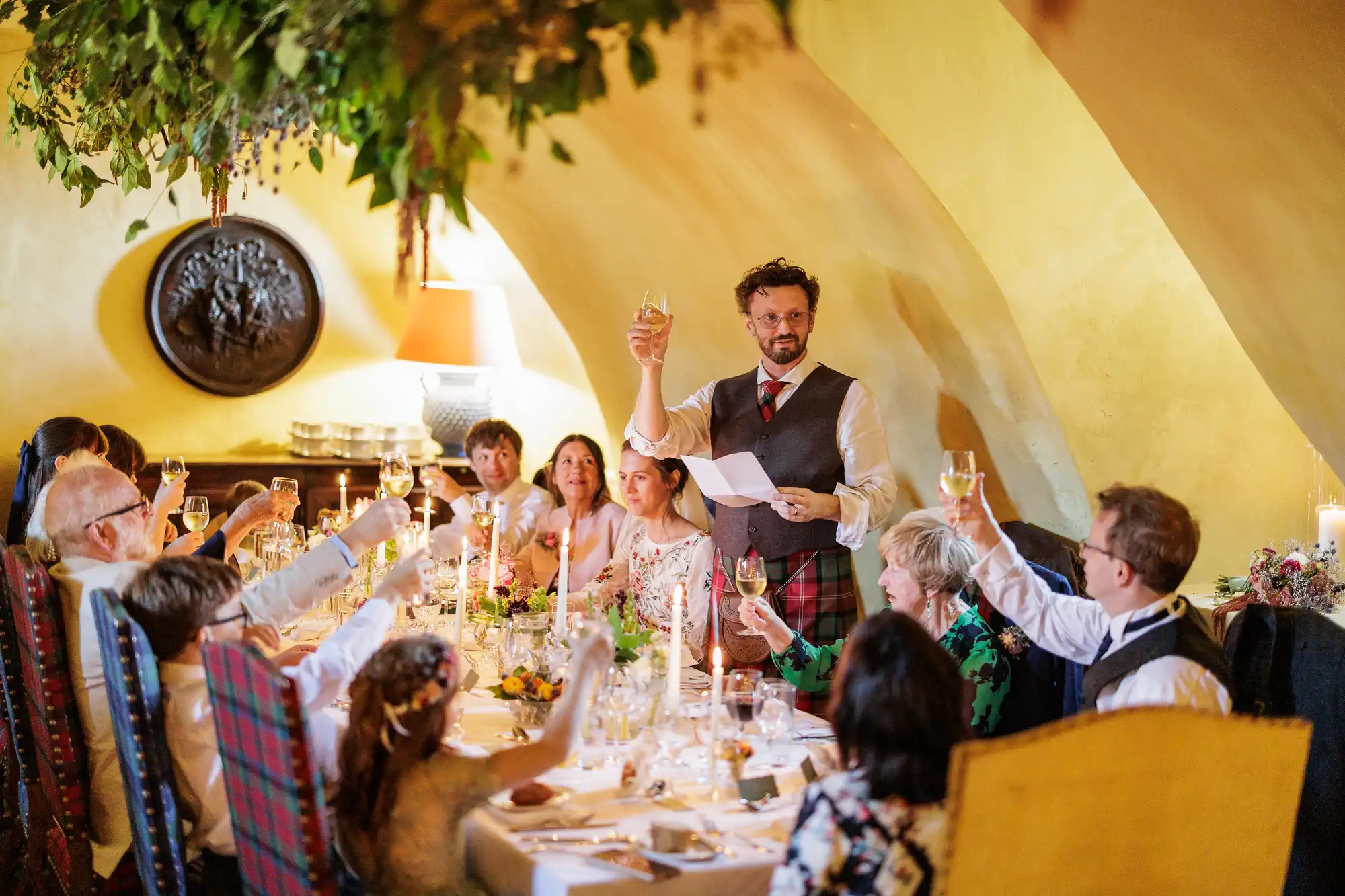 A group of people sit around a long, elegantly laid table in a warmly lit room. A man standing at the head of the table raises a glass in a toast, holding a sheet of paper. Guests smile and raise glasses, surrounded by floral arrangements and hanging greenery.