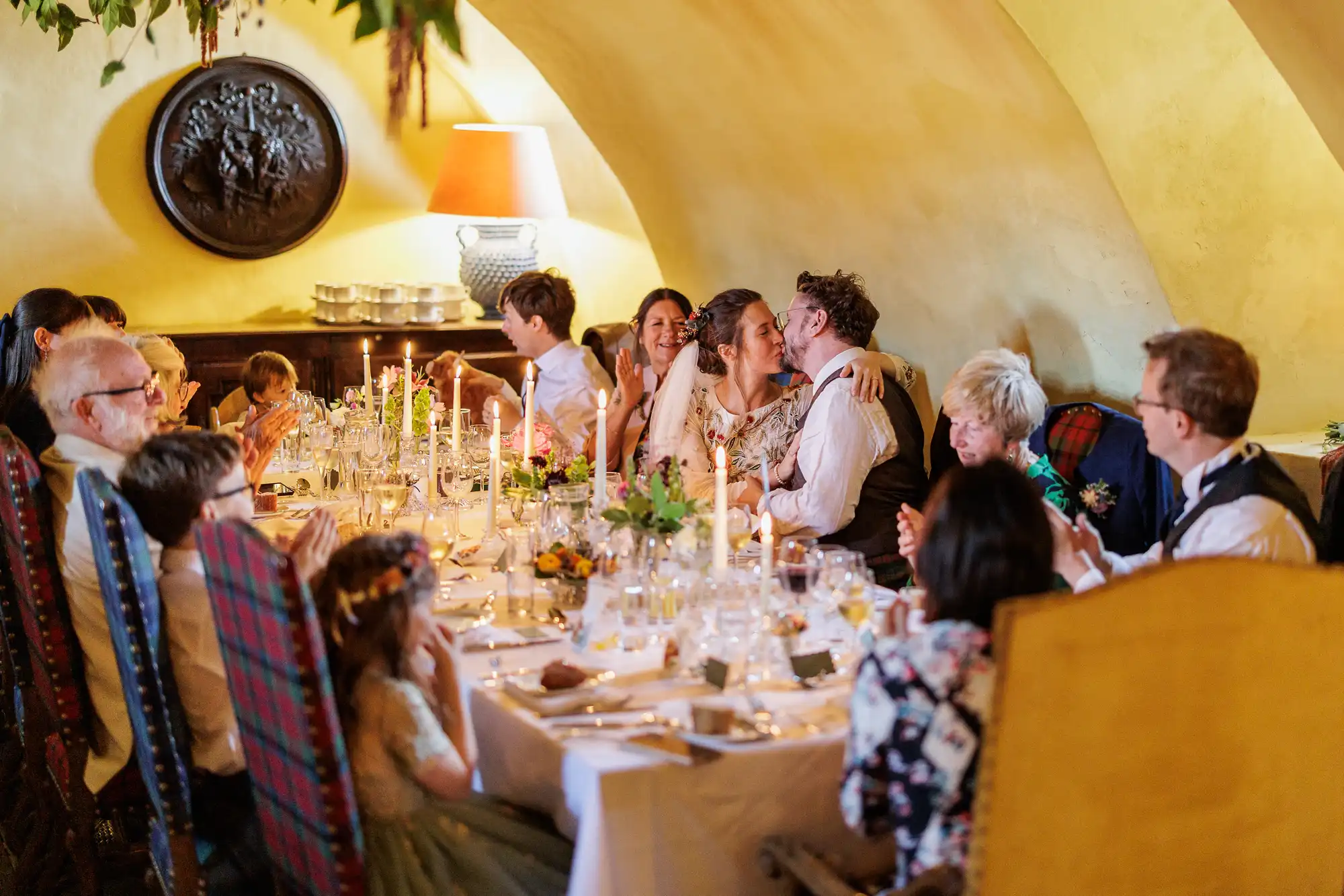 A wedding reception scene with guests seated at a long, candlelit table. A couple in the centre shares a kiss whilst others smile, clap, and watch. The room has warm lighting, yellow walls, and elegant decor, creating a joyful and intimate atmosphere.