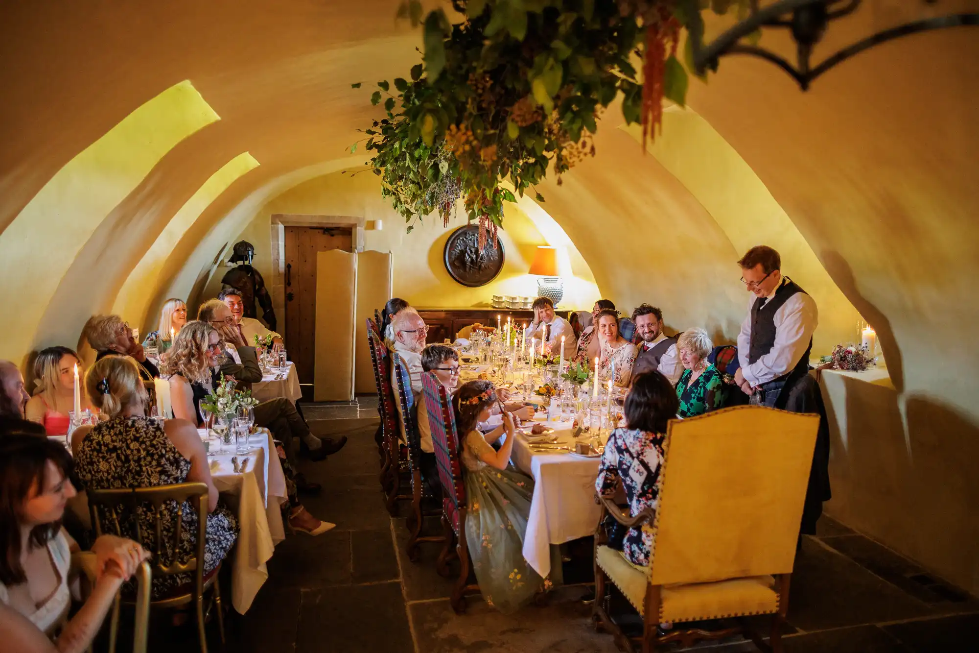 A group of people dressed formally are seated at long tables in a cosy, yellow-toned arched room with stone floors. The tables are decorated with flowers and candles. A waiter stands by, and greenery hangs from the ceiling. Everyone looks joyful, chatting and smiling.
