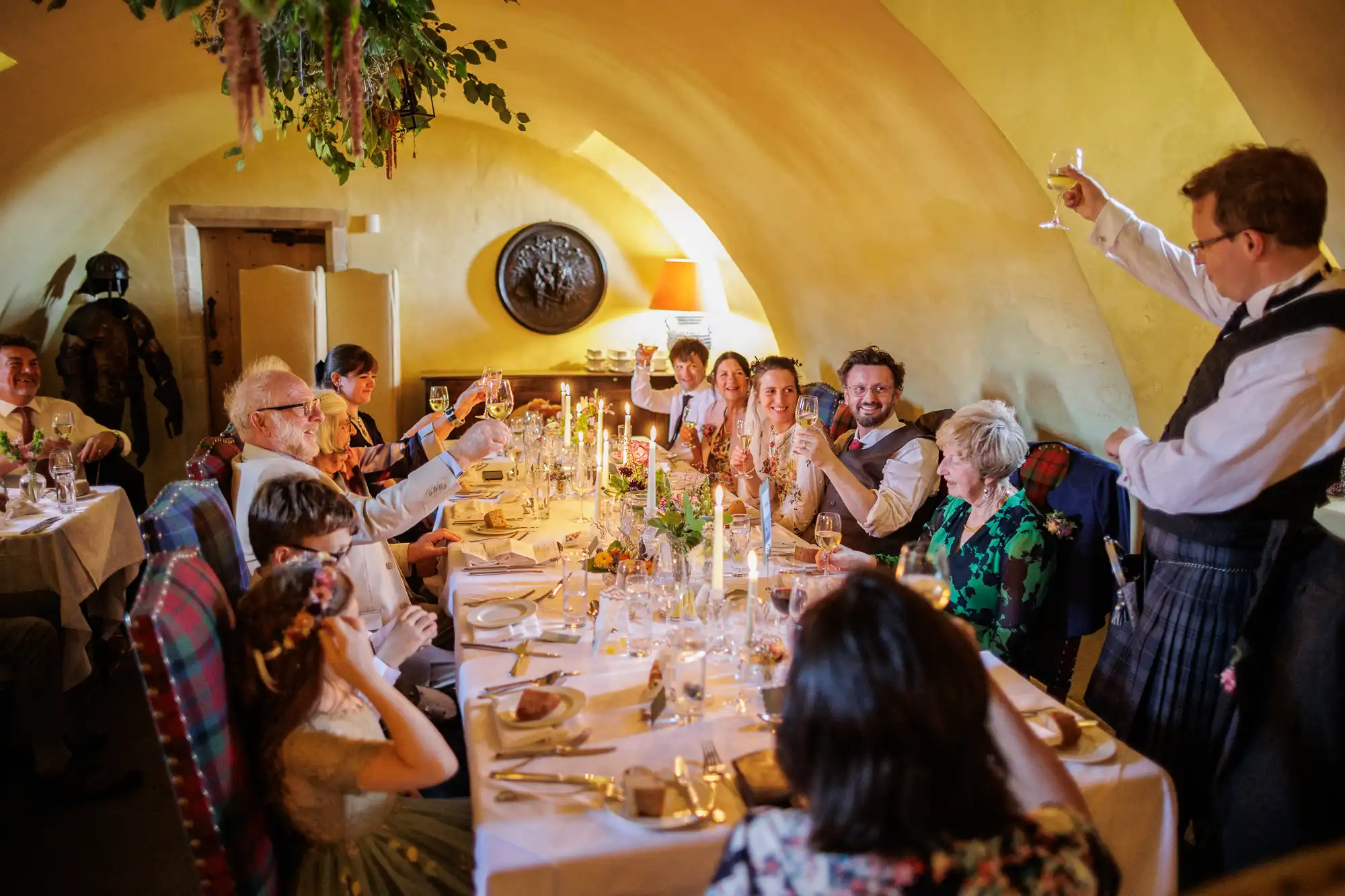 A lively group of people sit around a long, candlelit table in a warmly lit room with arched ceilings, smiling and raising glasses in a toast, celebrating at what appears to be a festive gathering or wedding reception.