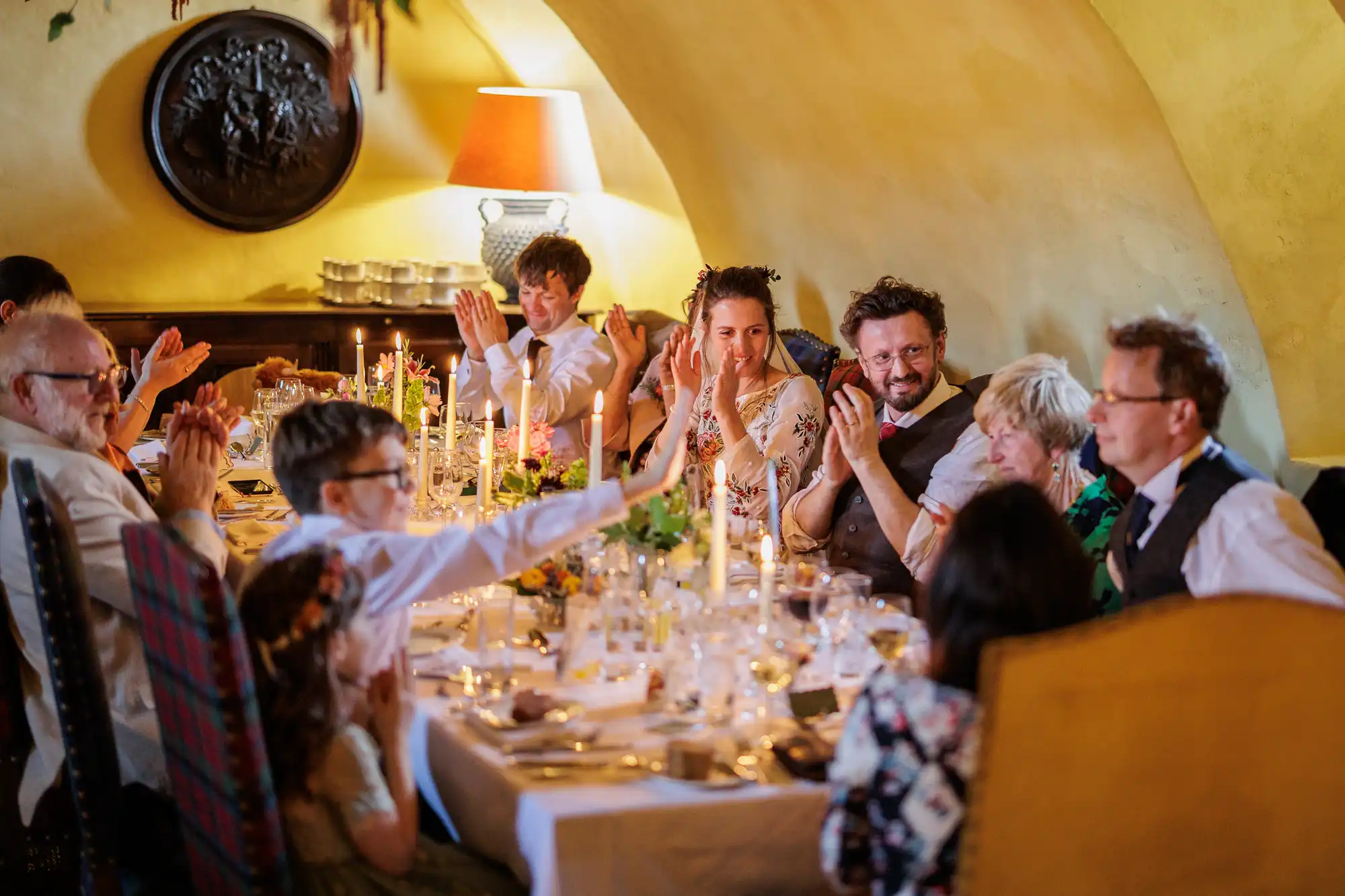 A group of people in formal attire sit around a candlelit dining table in a warm, cosy room. They are smiling, clapping, and engaging with each other, suggesting a celebratory event such as a wedding or family gathering.