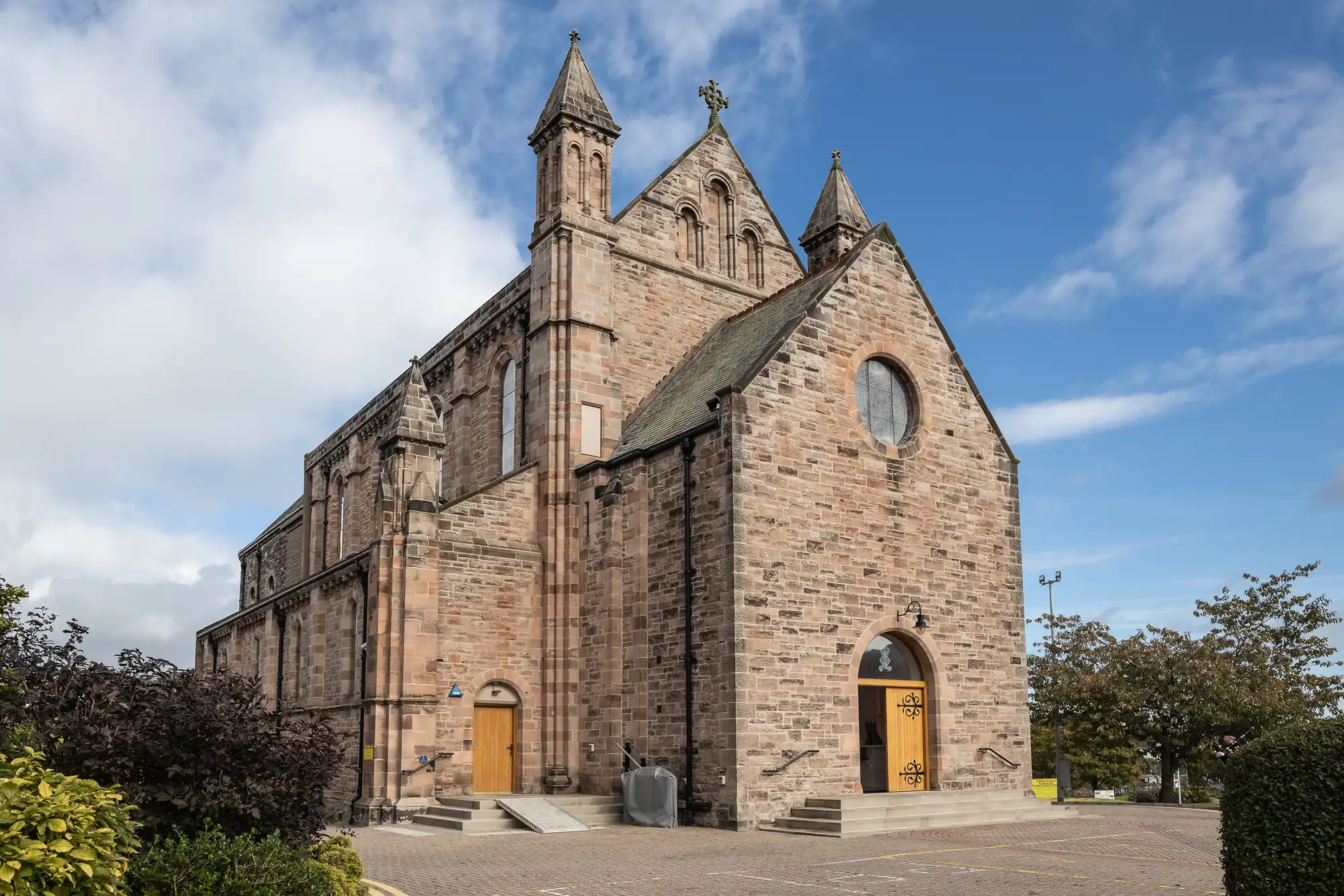 A large stone church with two pointed towers, arched windows, and three wooden doors. The building has a steep gabled roof and is surrounded by a paved area, trees, and bushes under a partly cloudy sky.
