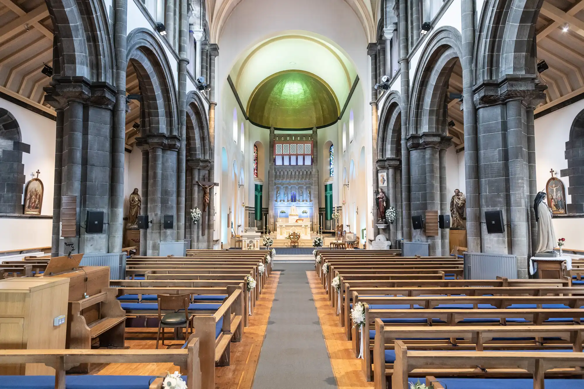 Wide-angle view of a grand church interior with stone arches, wooden pews, statues, religious paintings, and a central aisle leading to an ornate altar under a domed ceiling. The space is well-lit and decorated with white flowers along the pews.