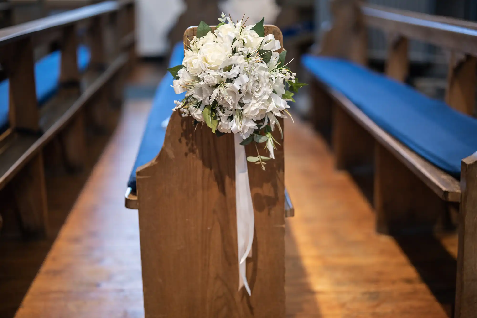 A close-up of a wooden church pew decorated with a bouquet of white flowers and greenery, tied with a white ribbon. The pews have blue cushions and are arranged in rows on a polished wooden floor. The background is softly blurred.
