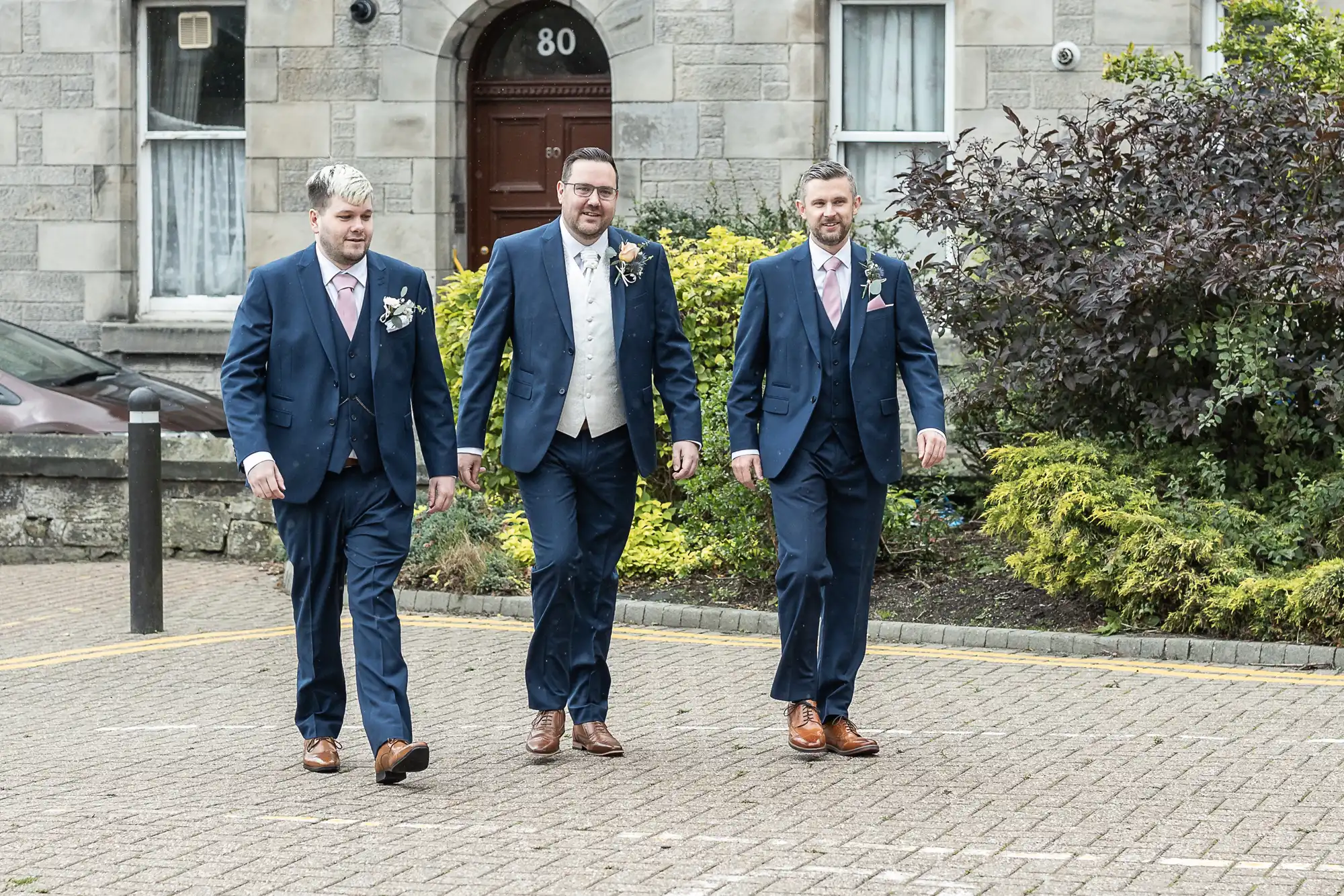 Three men in blue suits with pink ties and buttonholes walk together on a paved path in front of a stone building with a brown door marked “80.” Green shrubs and small trees line the walkway behind them. All appear dressed for a formal occasion.