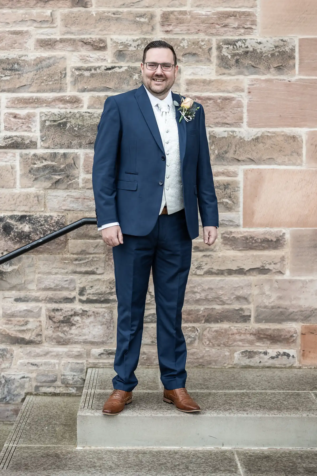 A man stands smiling in front of a stone wall, wearing a blue suit with a white waistcoat, white shirt, and brown shoes. He has short dark hair, glasses, and a buttonhole pinned to his lapel. His hands rest at his sides.