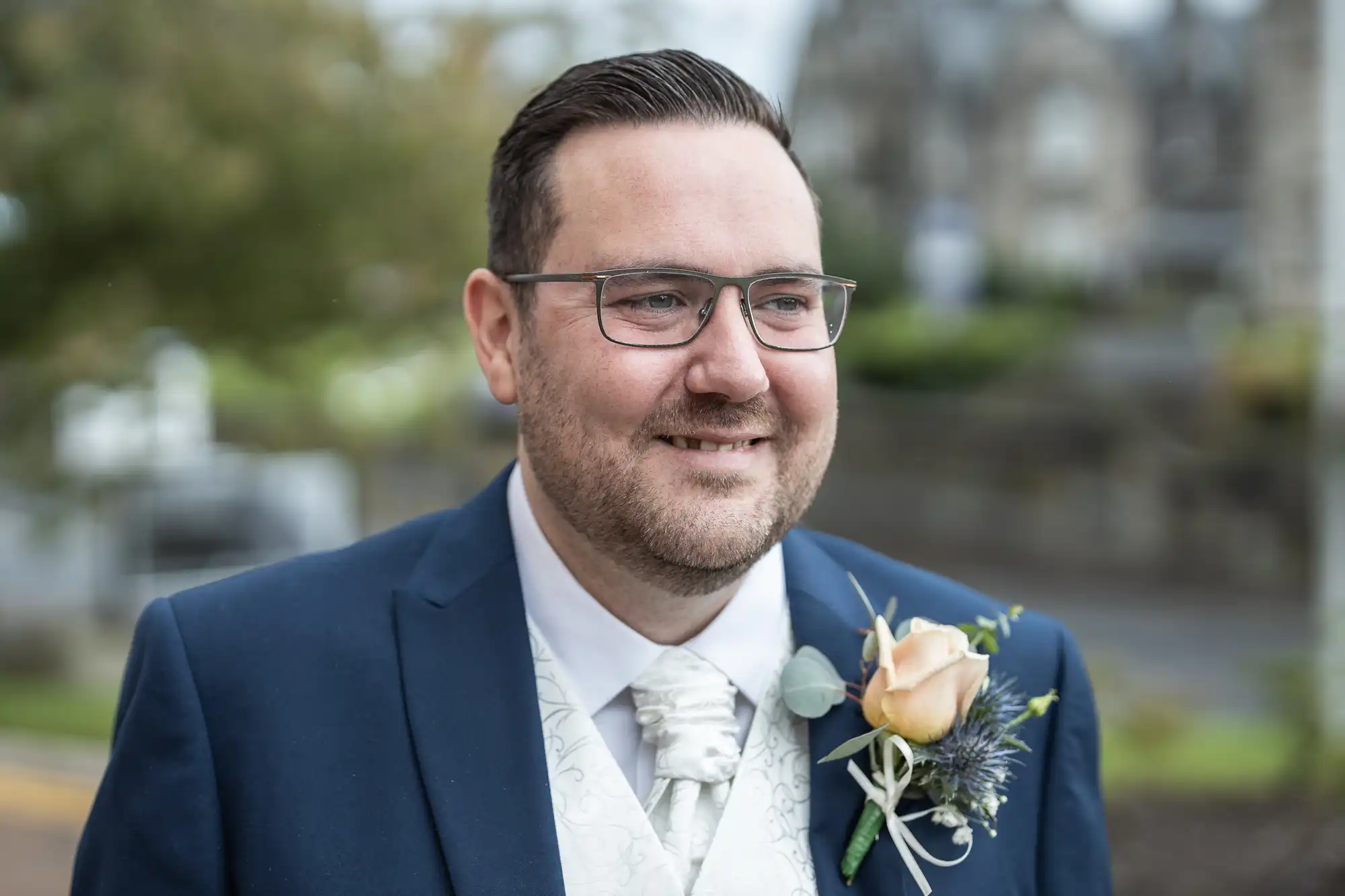 A man with short dark hair, glasses, and light stubble smiles whilst wearing a navy blue suit with a white waistcoat and tie. He has a peach rose buttonhole pinned to his lapel. The blurred outdoor background suggests a formal event.