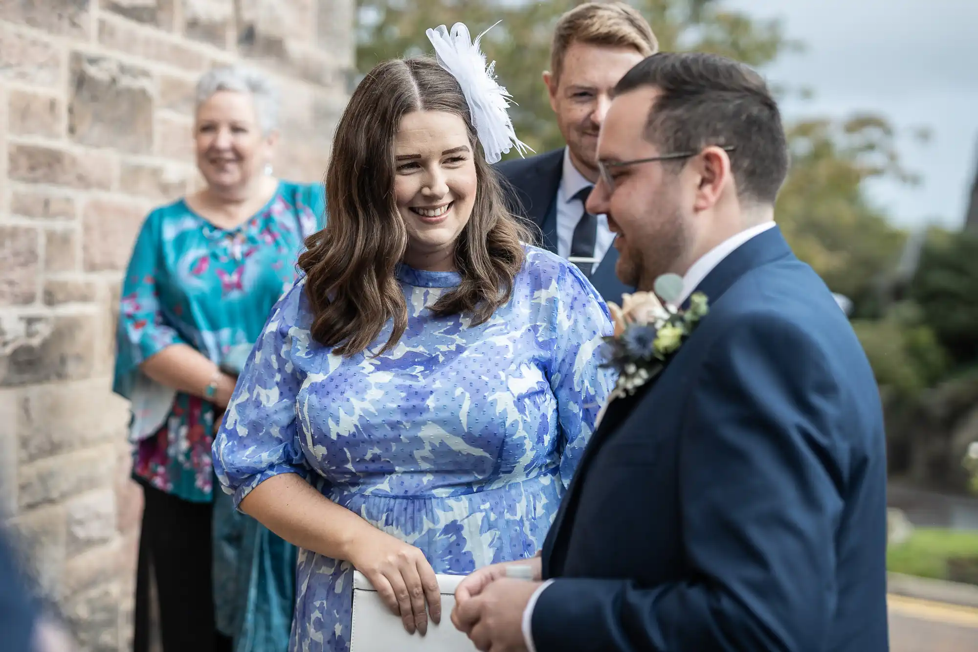 A woman in a light blue floral dress and white fascinator smiles at a man in a dark suit and glasses. Two other people, one in a blue-green dress and another in a suit, stand behind them outside near a stone wall. It appears to be a joyful occasion.