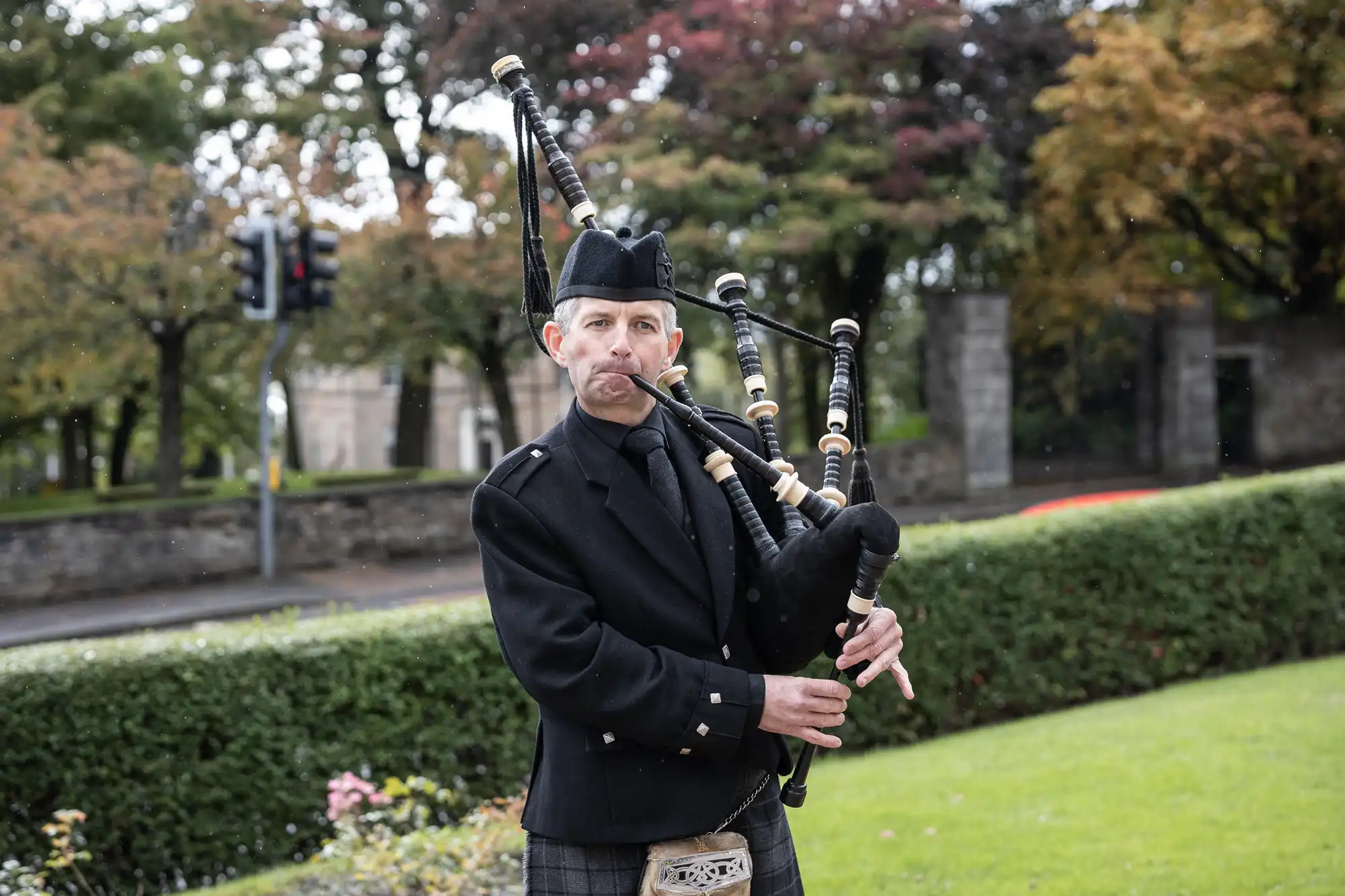 A man wearing traditional Scottish attire, including a black jacket, hat, and tartan clothing, plays bagpipes outdoors. He stands on grass near a hedge, with trees and stone walls in the background under cloudy skies.