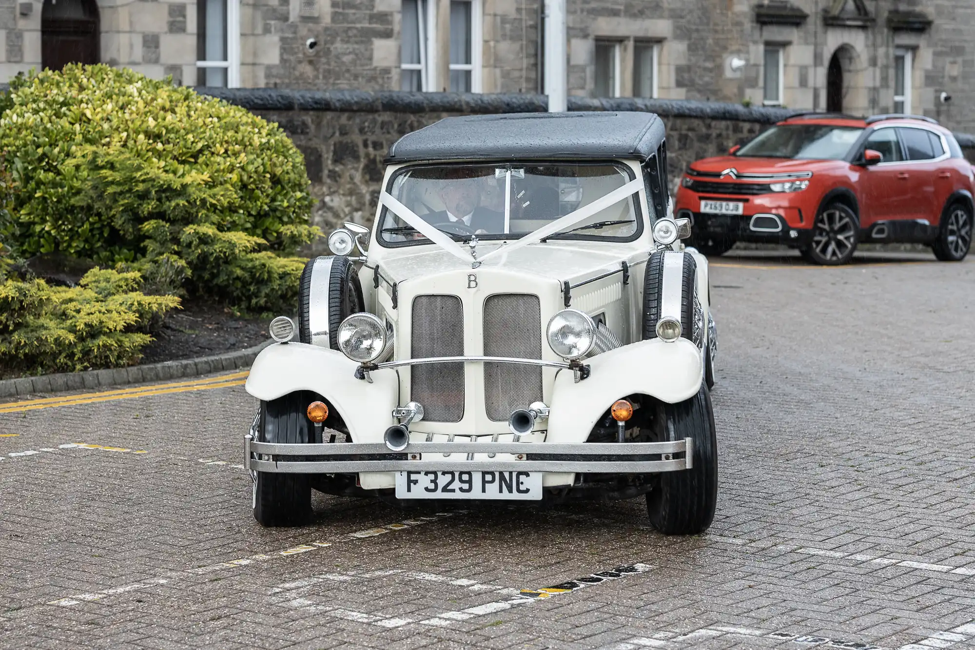 A vintage white convertible car with a black soft top, circular headlamps, and chrome details is parked on a wet street near green bushes. A red modern SUV and stone buildings are visible in the background. The car has a UK number plate: F329 PNE.